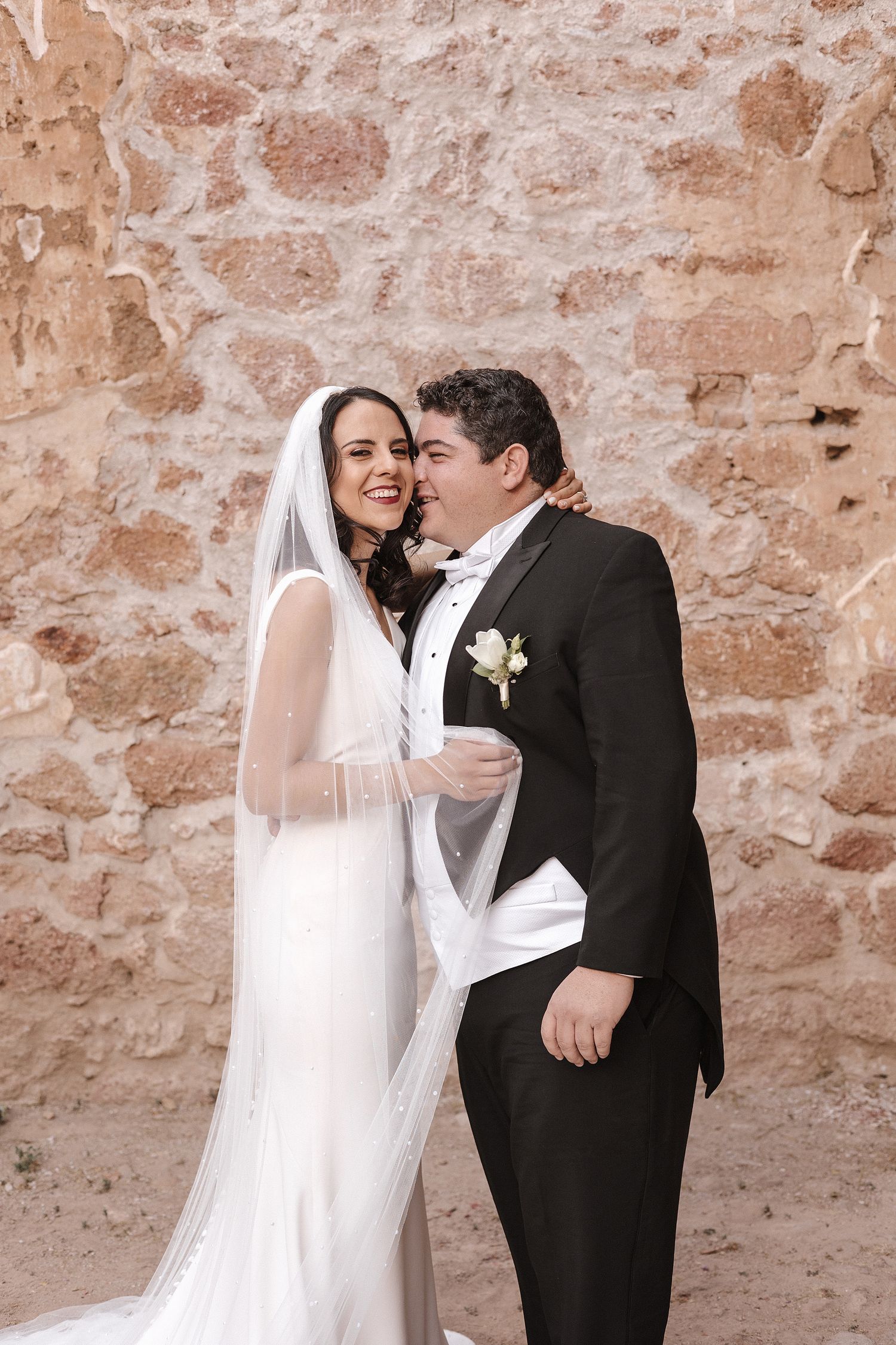 Joyful wedding couple embracing against a textured stone wall background.