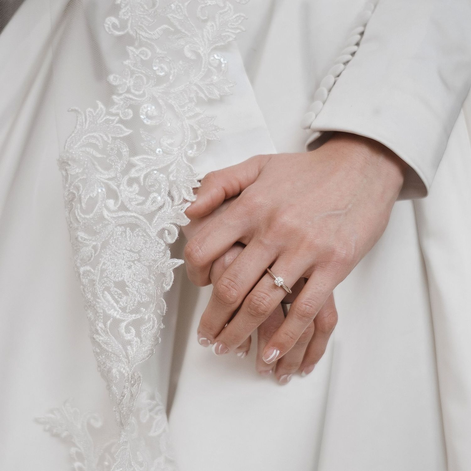 Close up of hands wearing wedding rings against white lace dress fabric.