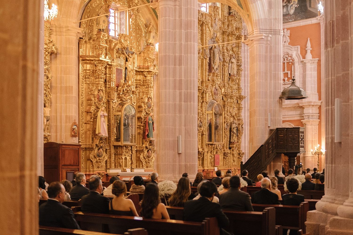 Wedding ceremony in an ornate cathedral with golden altars and guests seated in wooden pews.
