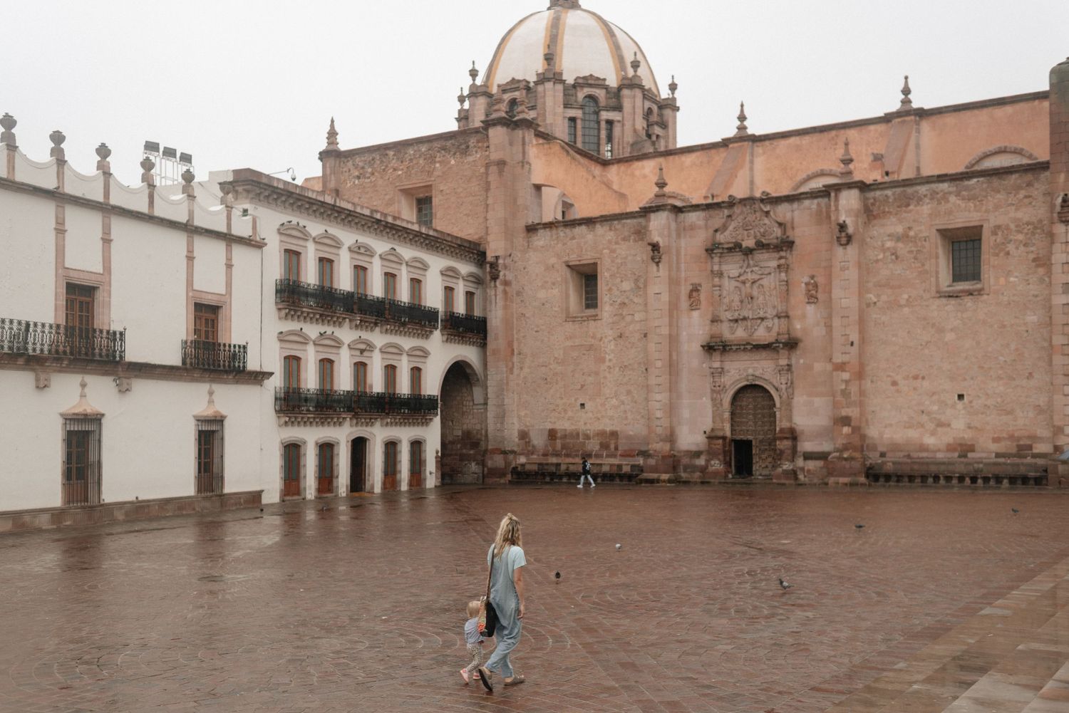 Person in blue poncho crosses rainy colonial plaza in front of historic Mexican church and Spanish colonial architecture.