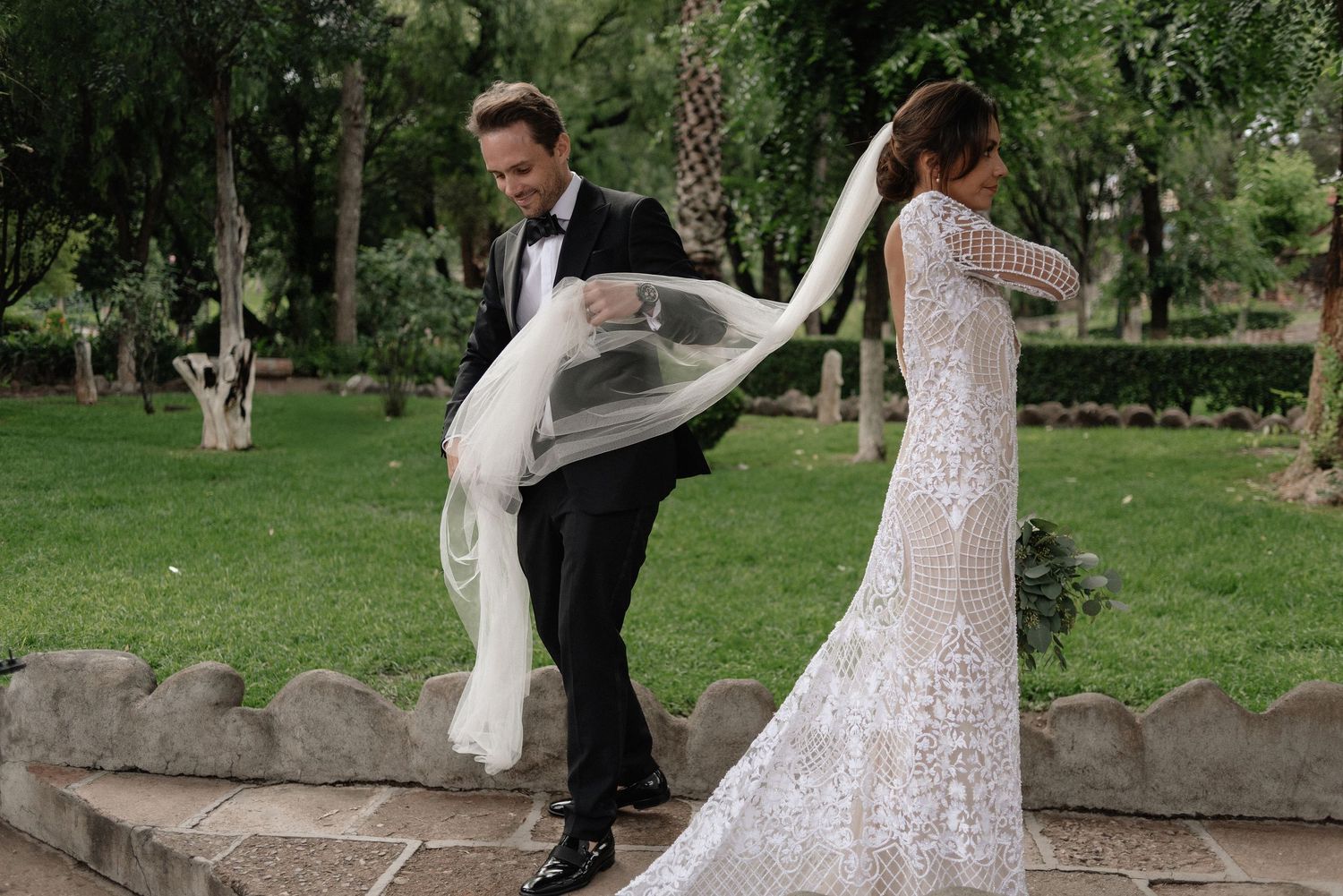 Two people in wedding attire playfully pose together in a garden setting with flowing white dress fabric caught in motion.