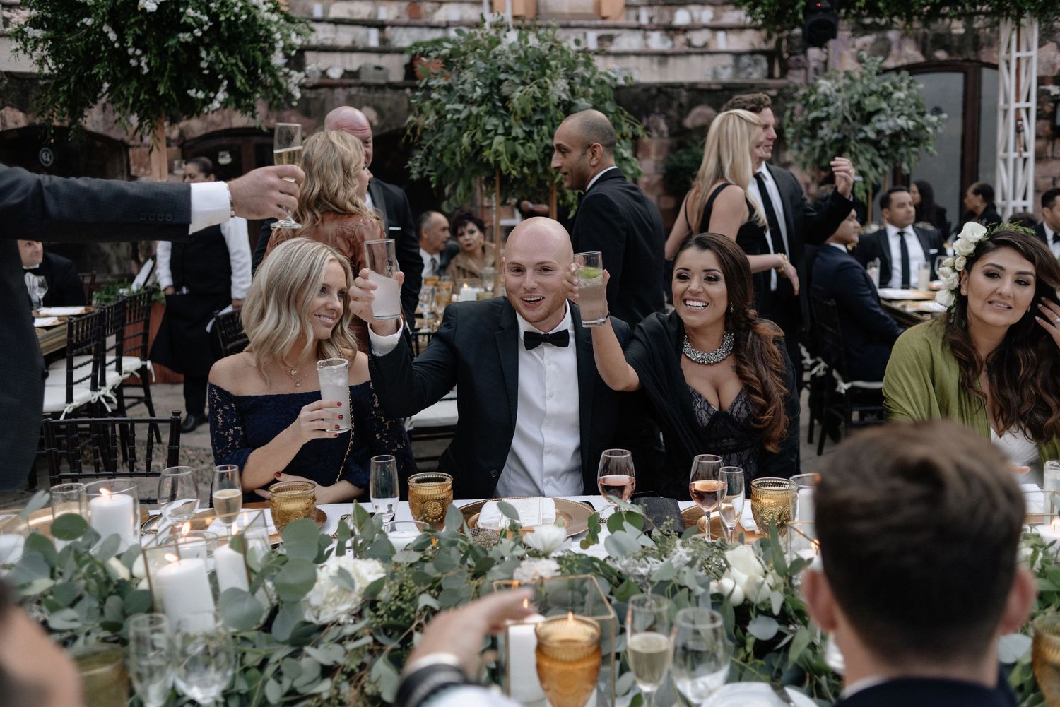 Wedding guests enjoy lively conversation at an elegant outdoor reception with candlelit table settings and greenery decor.
