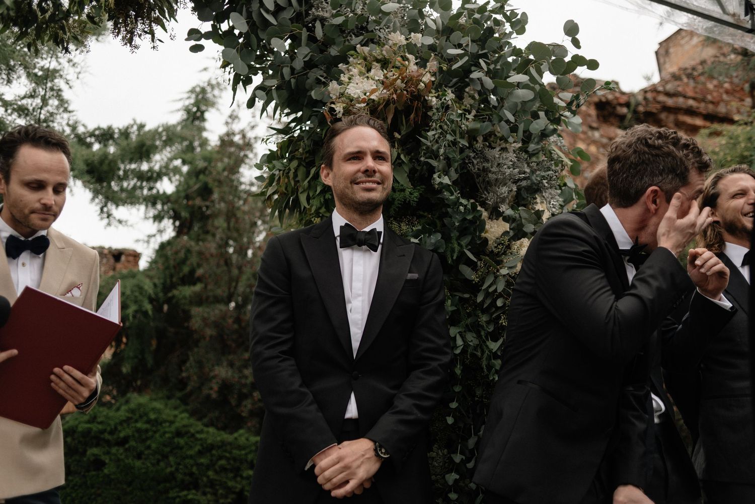 At an outdoor wedding ceremony, emotional reactions are captured with formal attire and a floral arch in the background.