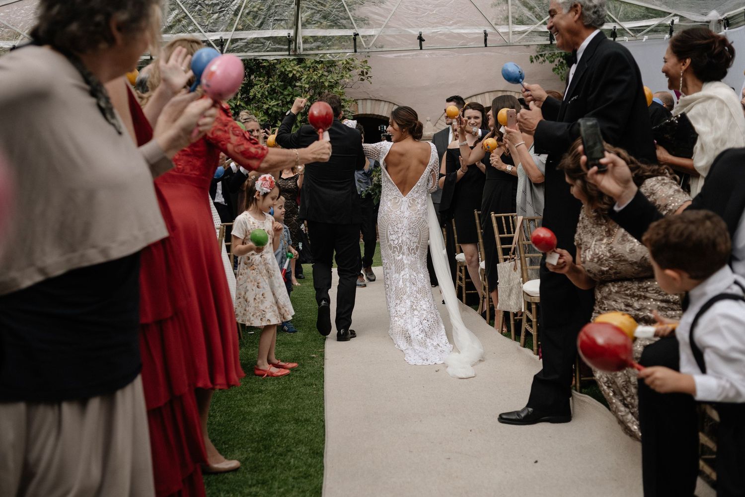 Wedding guests blow bubbles along the aisle during an outdoor garden ceremony as newlyweds walk past in celebration.