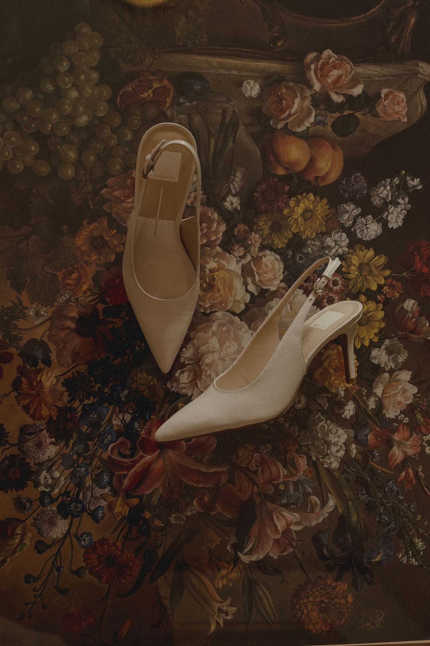White pointed-toe slingback heels with ankle straps displayed against a dark floral vintage painting background.