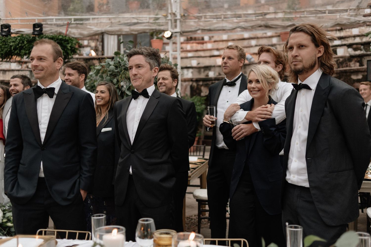 A line of groomsmen in black tuxedos stand together at an outdoor reception venue with exposed brick walls.