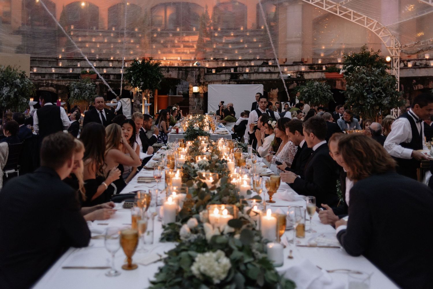 An elegant wedding reception dinner with long tables, candlelight and greenery in an industrial-style venue.
