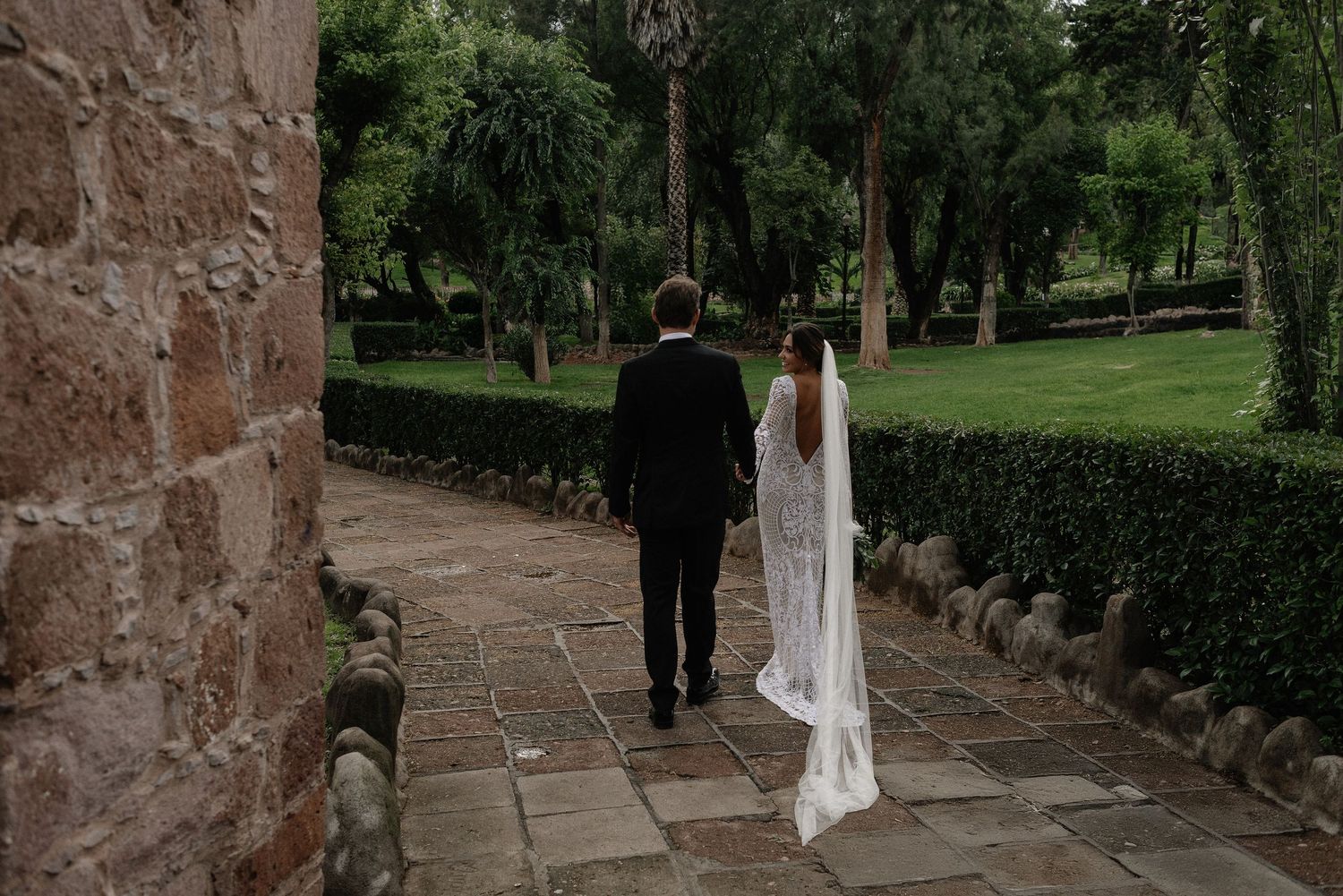 A couple in wedding attire walks down a stone path bordered by lush gardens and greenery at a historic venue.