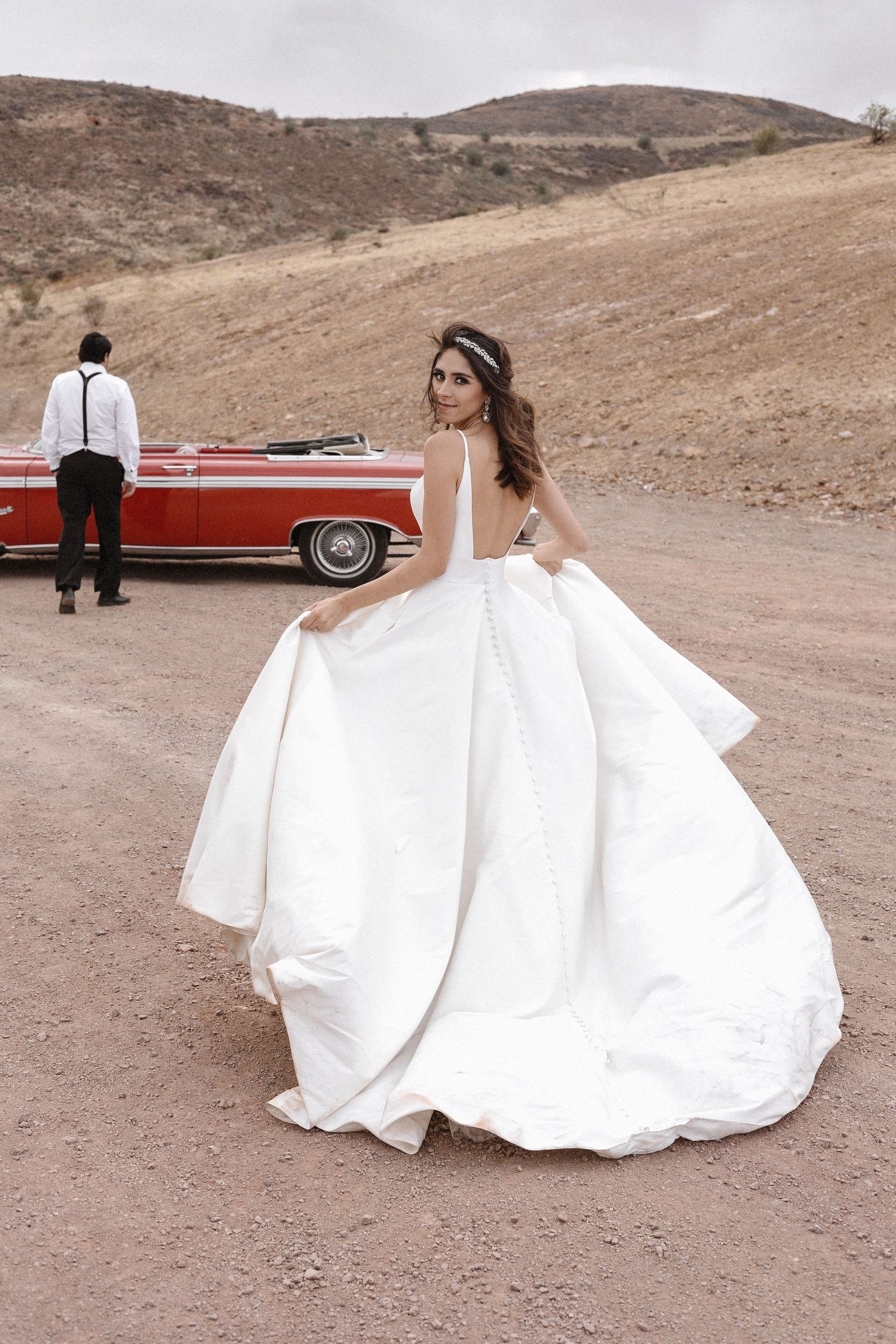 A bride in a flowing white ballgown poses dramatically in front of a red vintage car in a desert landscape.