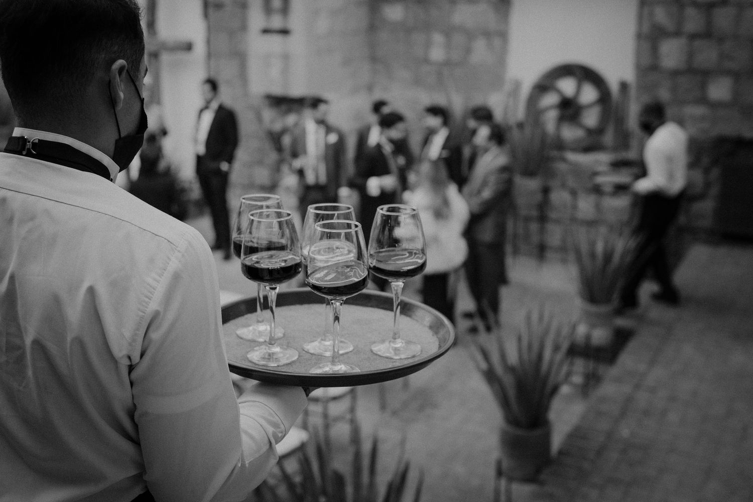 Wedding reception guests mingle outdoors while servers carry trays of drinks at dusk.