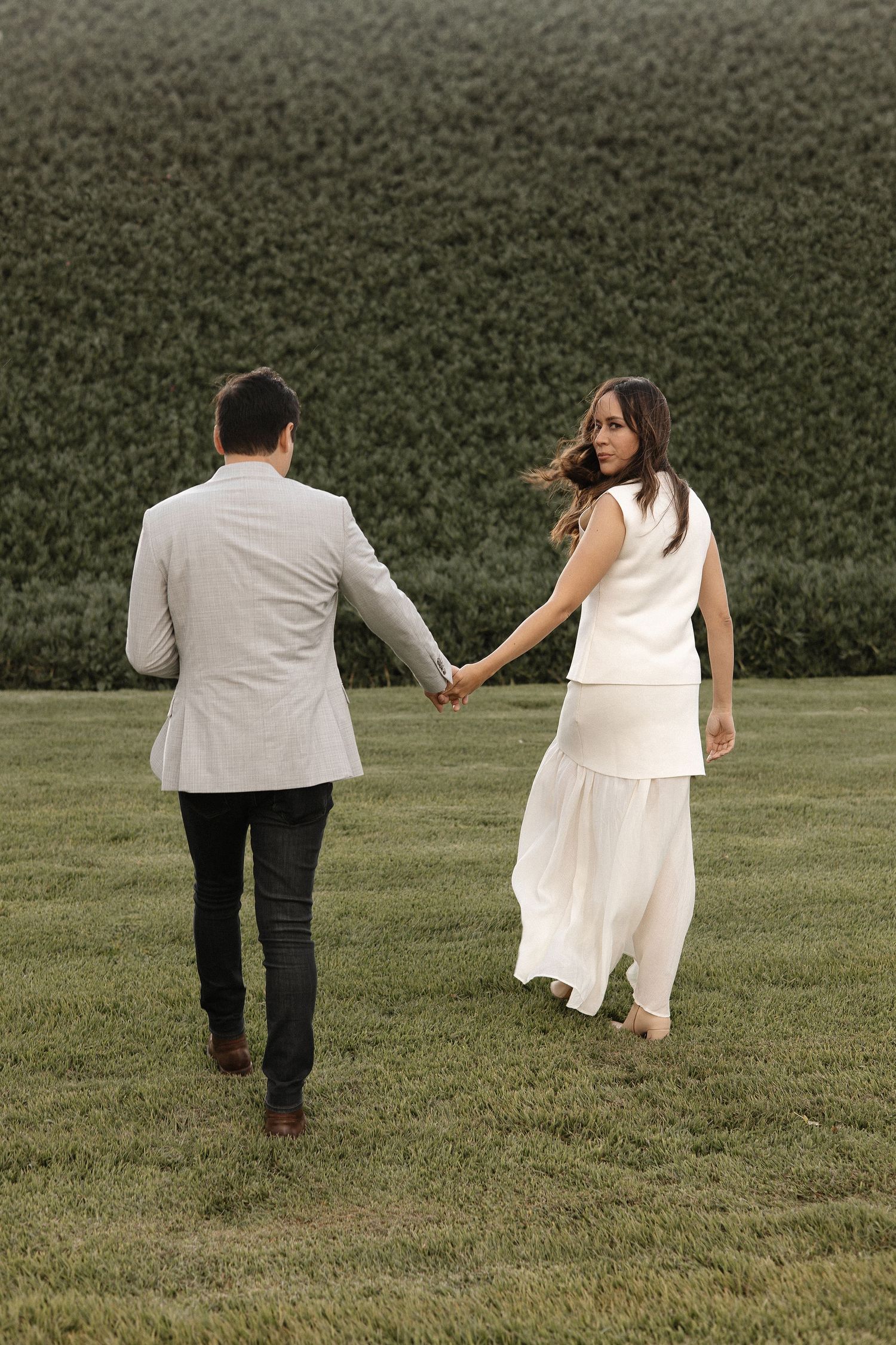 A couple walks hand in hand across a grassy field with a lush green backdrop on their wedding day.