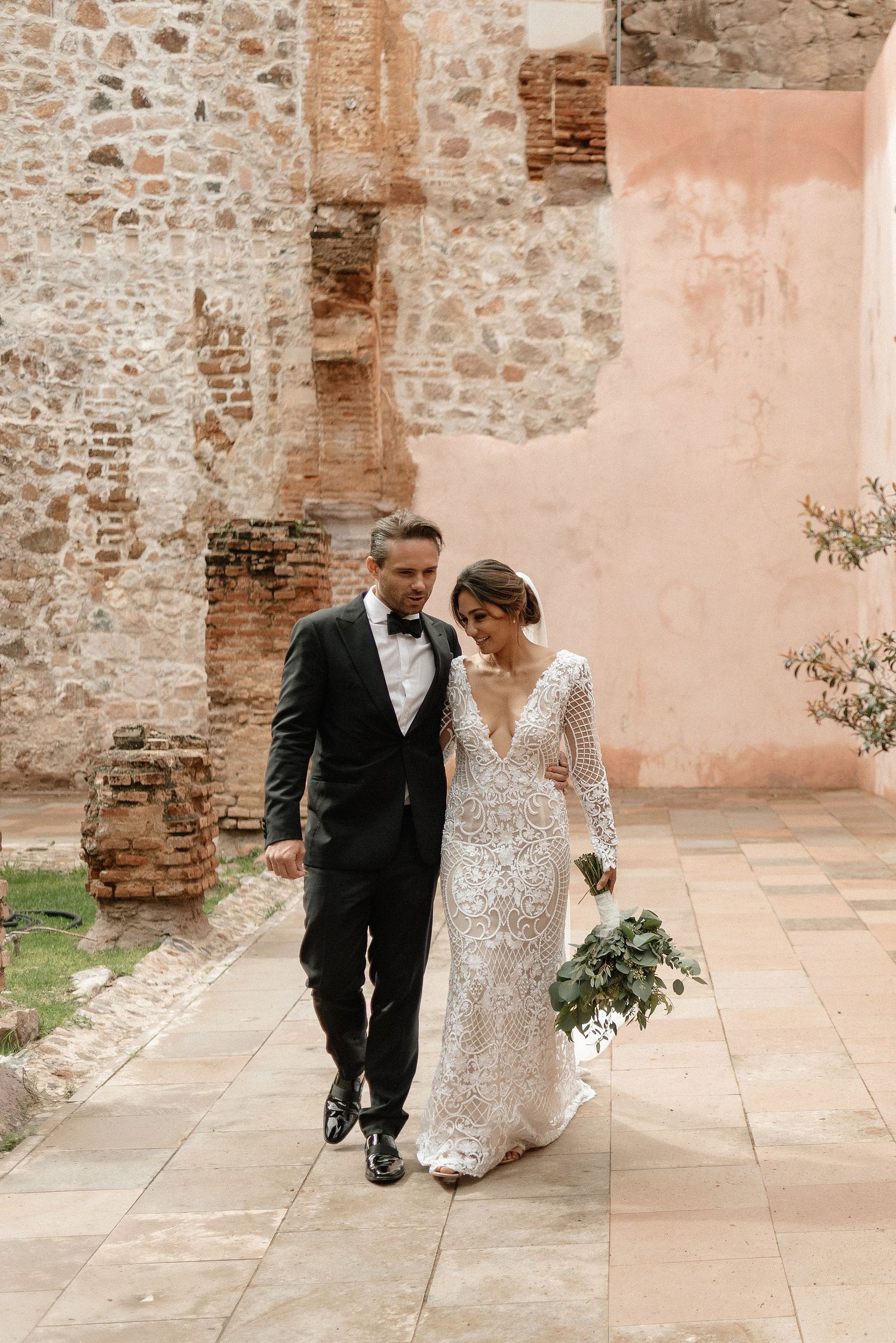 Elegant couple walking together at rustic stone venue with pink walls, dressed in formal black tuxedo and lace wedding gown.