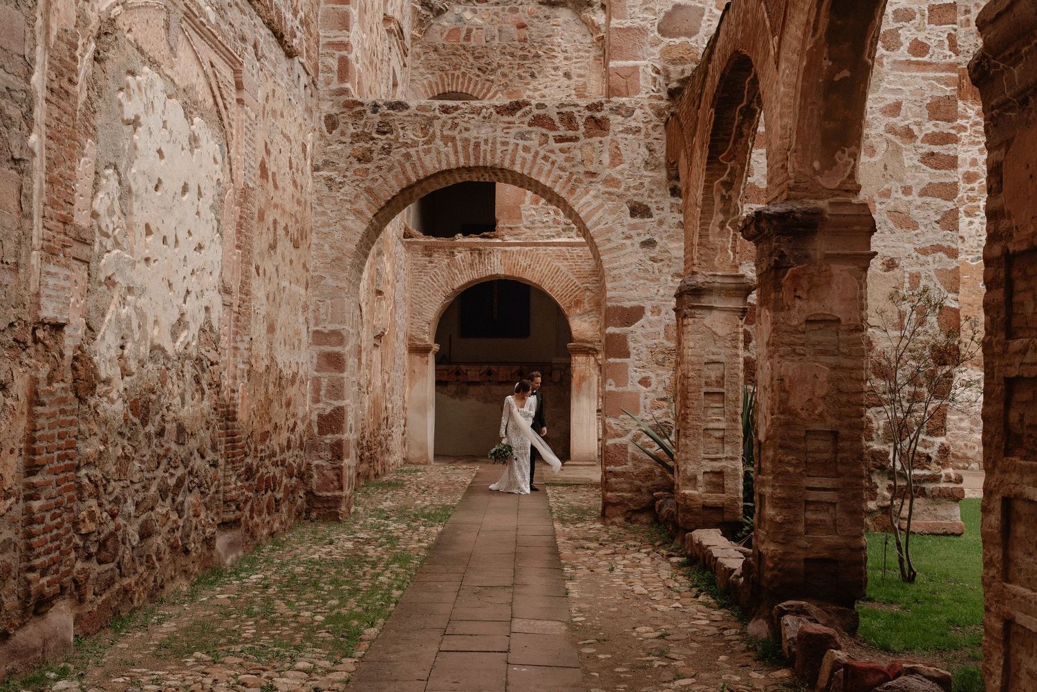 Warm-toned photograph of a figure in white standing beneath ancient brick archways in a historic ruin.