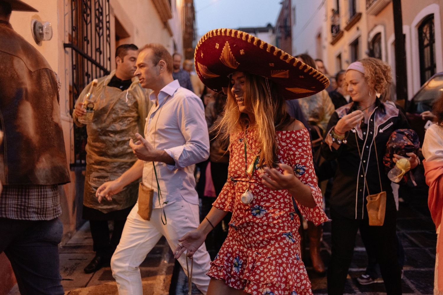 People celebrating in the streets of Mexico with someone wearing a traditional red sombrero and floral dress.