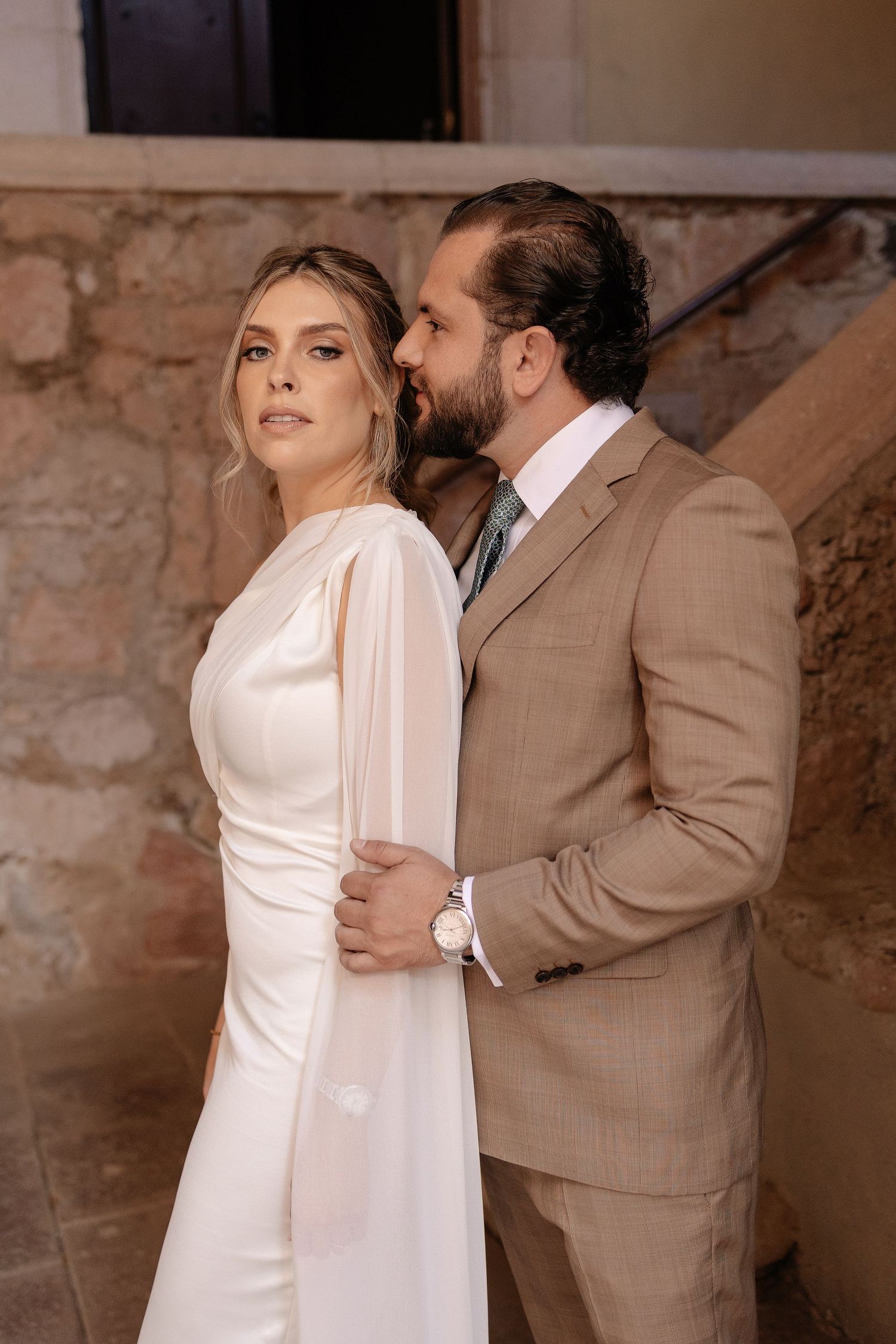 A romantic wedding couple poses against a stone wall, with the bride in white and groom in tan suit.