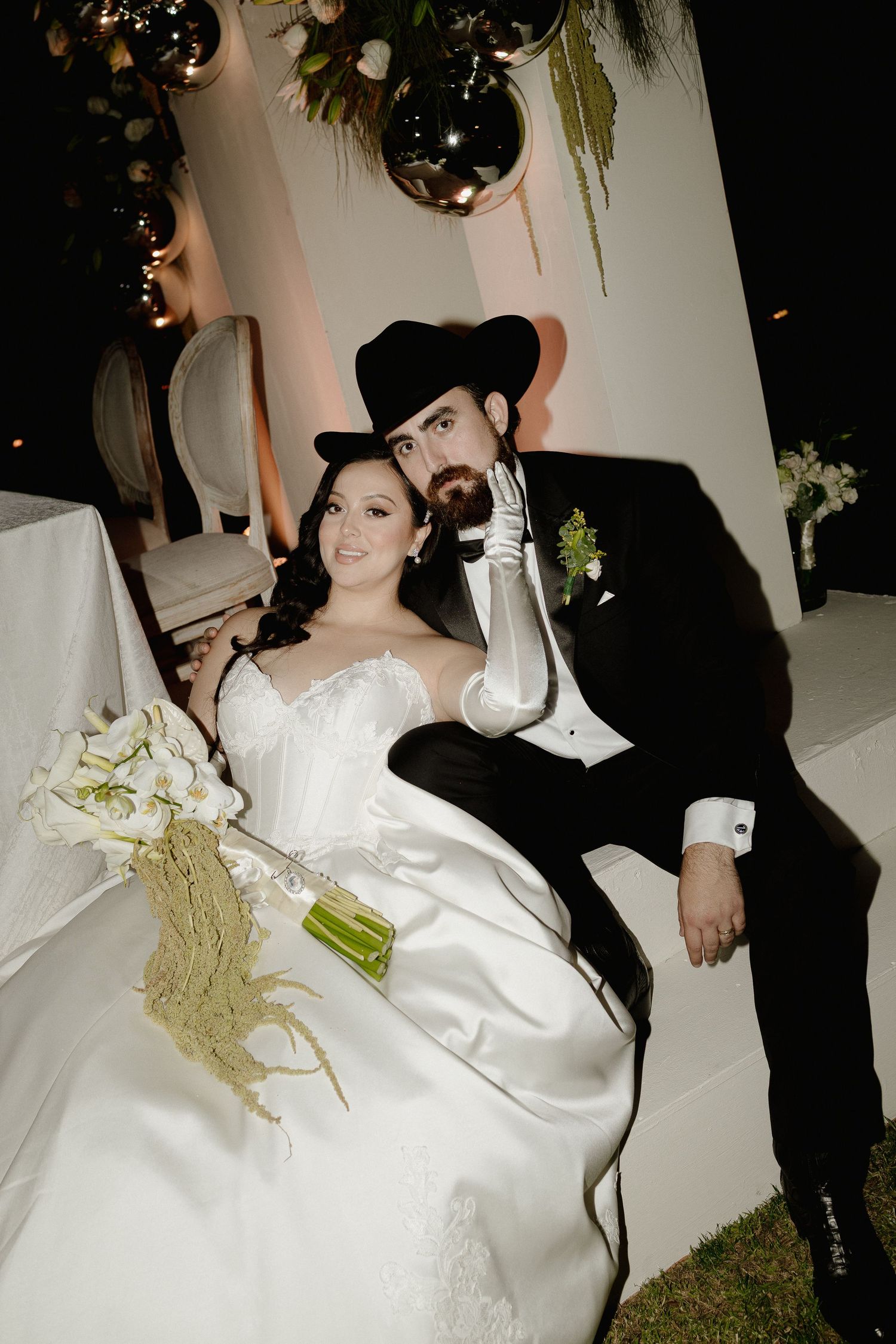 A couple in wedding attire poses together at their reception with silver disco balls and greenery decorations overhead.