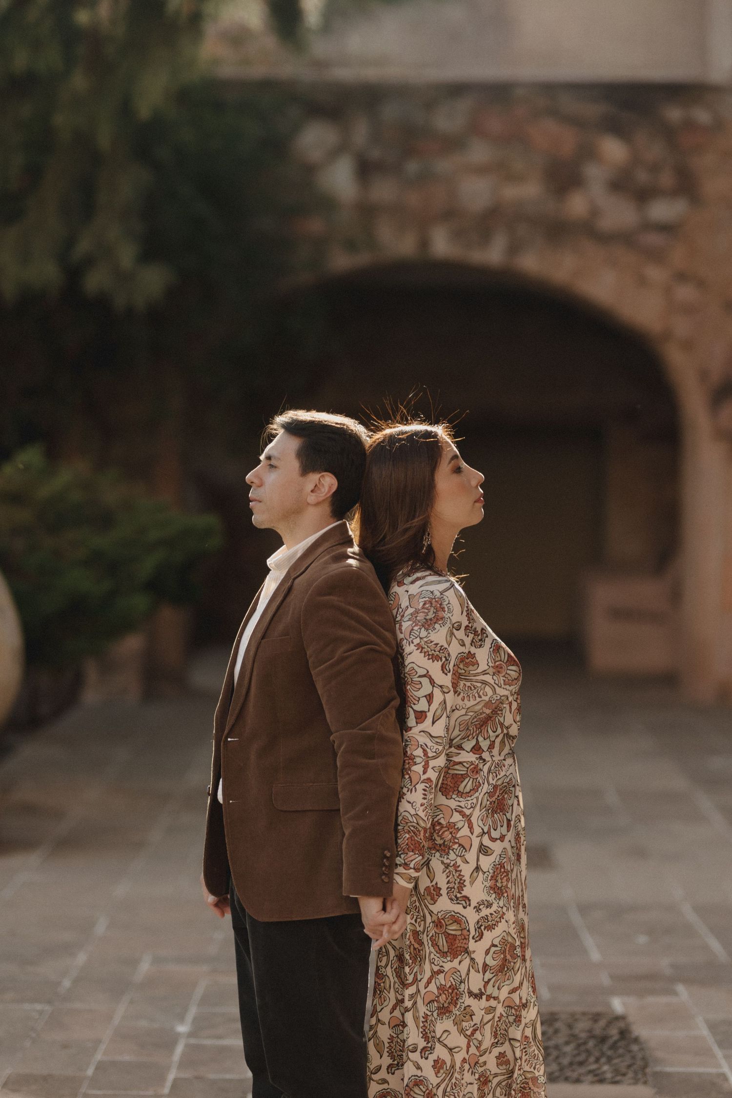 Couple in vintage attire poses back-to-back in a rustic courtyard setting with stone walls and natural lighting.