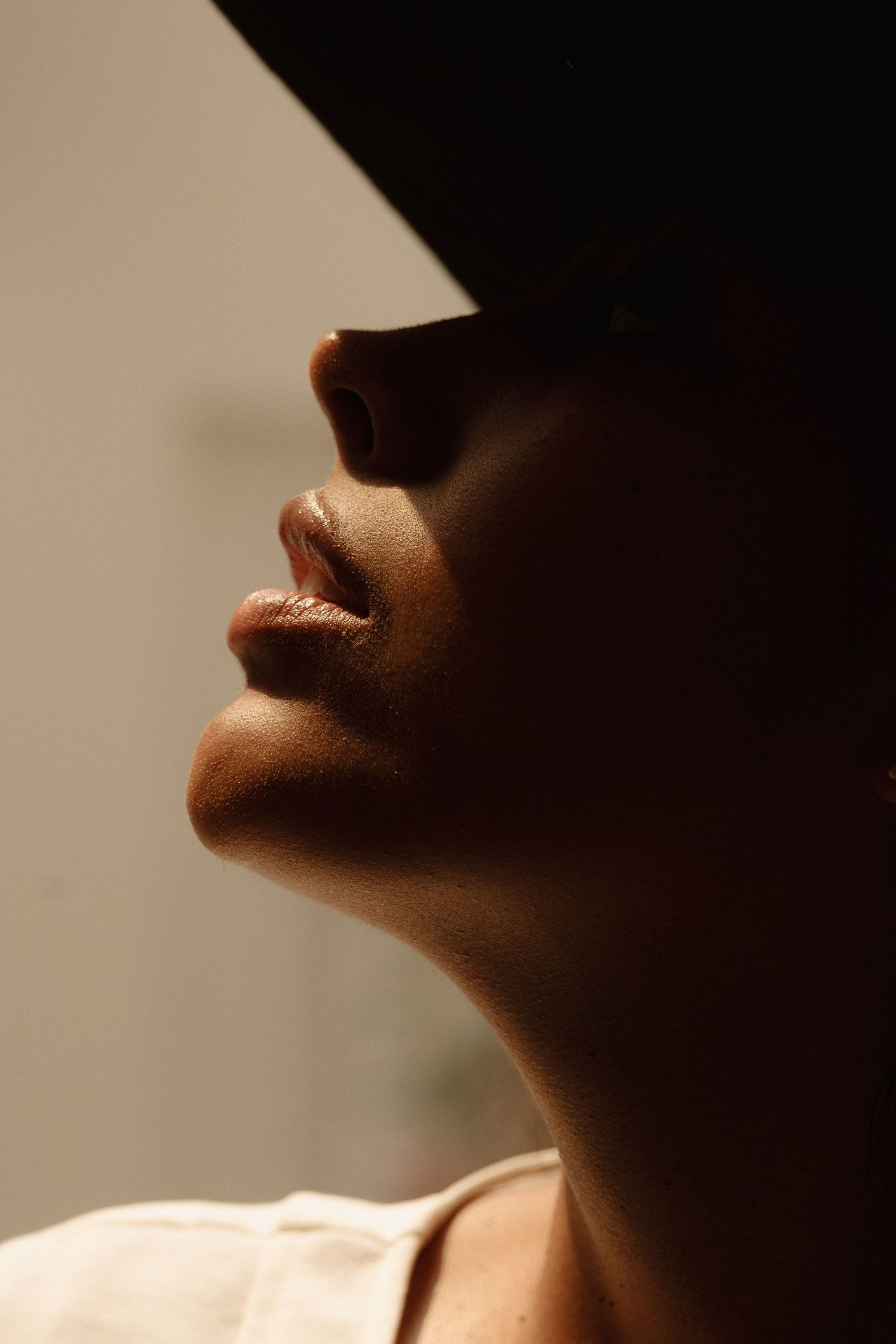 Close-up profile of a person wearing a dark hat, dramatic shadow lighting highlighting lips, nose, and neck.