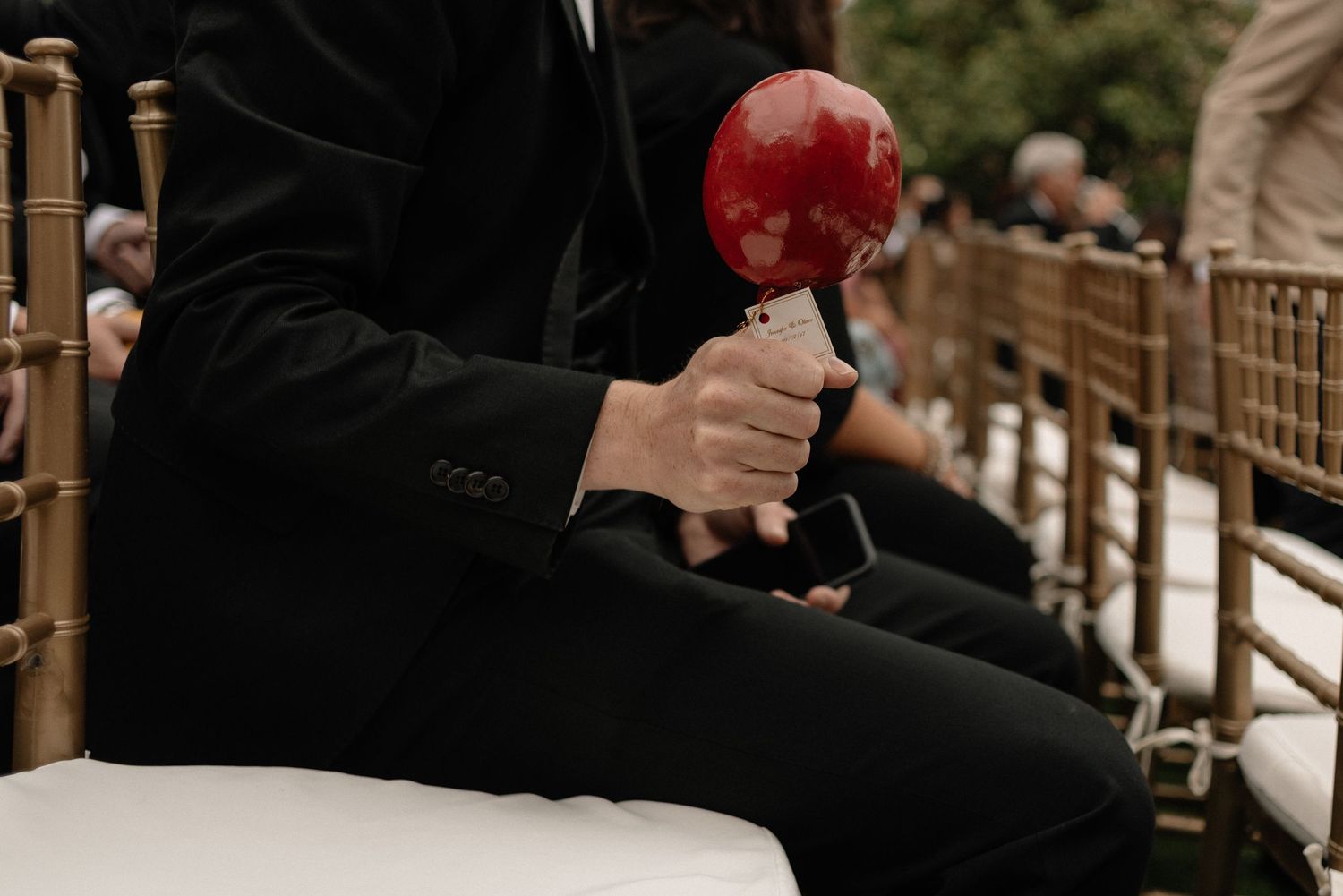 Someone wearing black formal attire holds a bright red candied apple at an outdoor winter event.