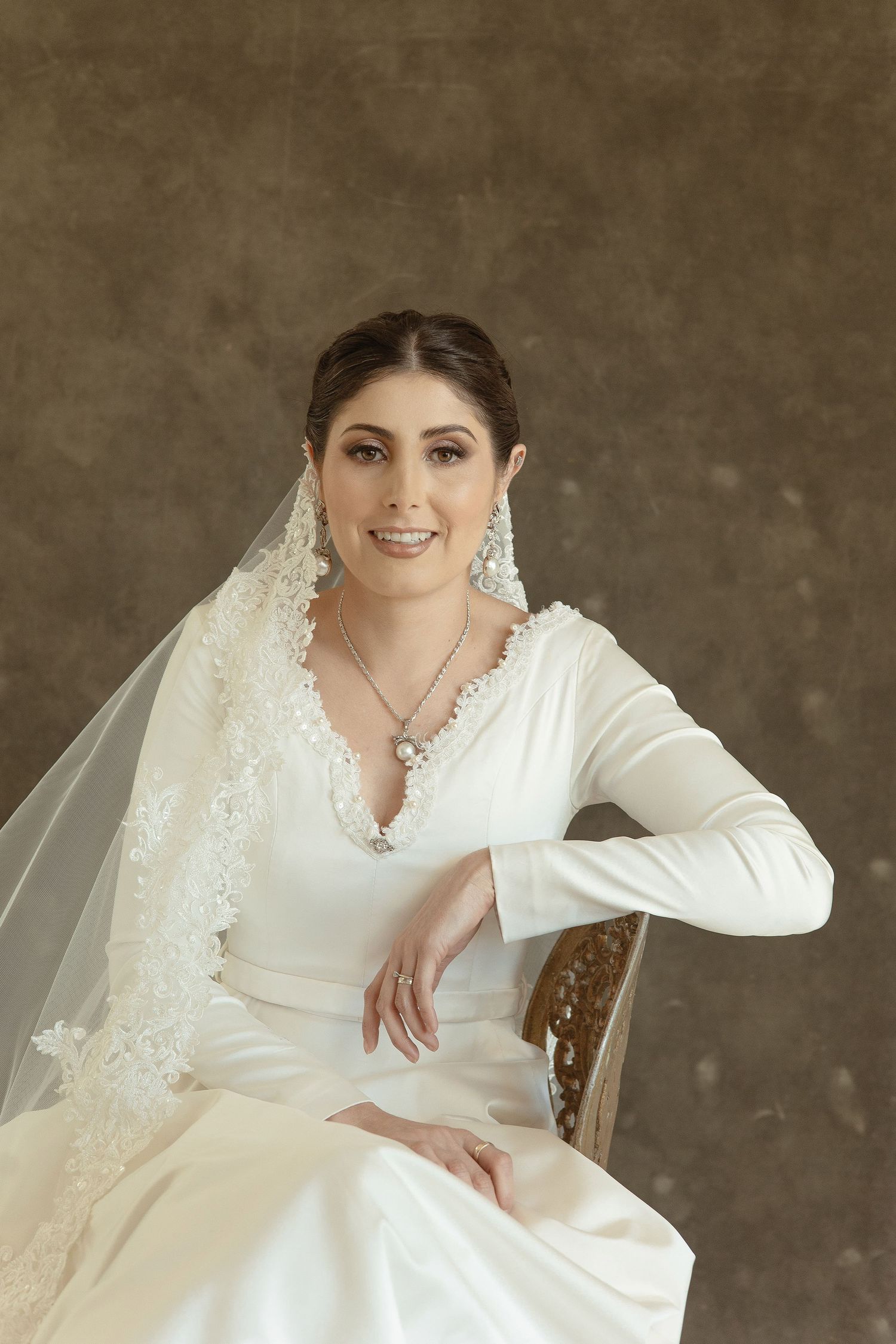 A bride in a classic long-sleeved white wedding dress and veil poses elegantly on an antique chair.