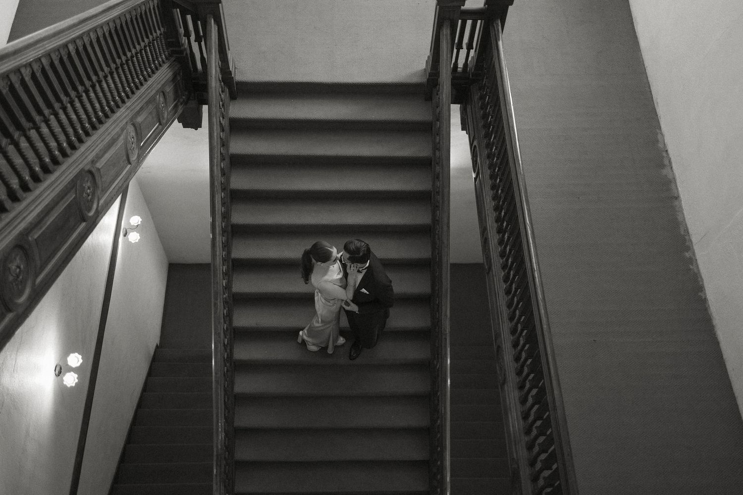A black and white aerial view of a couple dancing intimately on a grand staircase between ornate banisters.