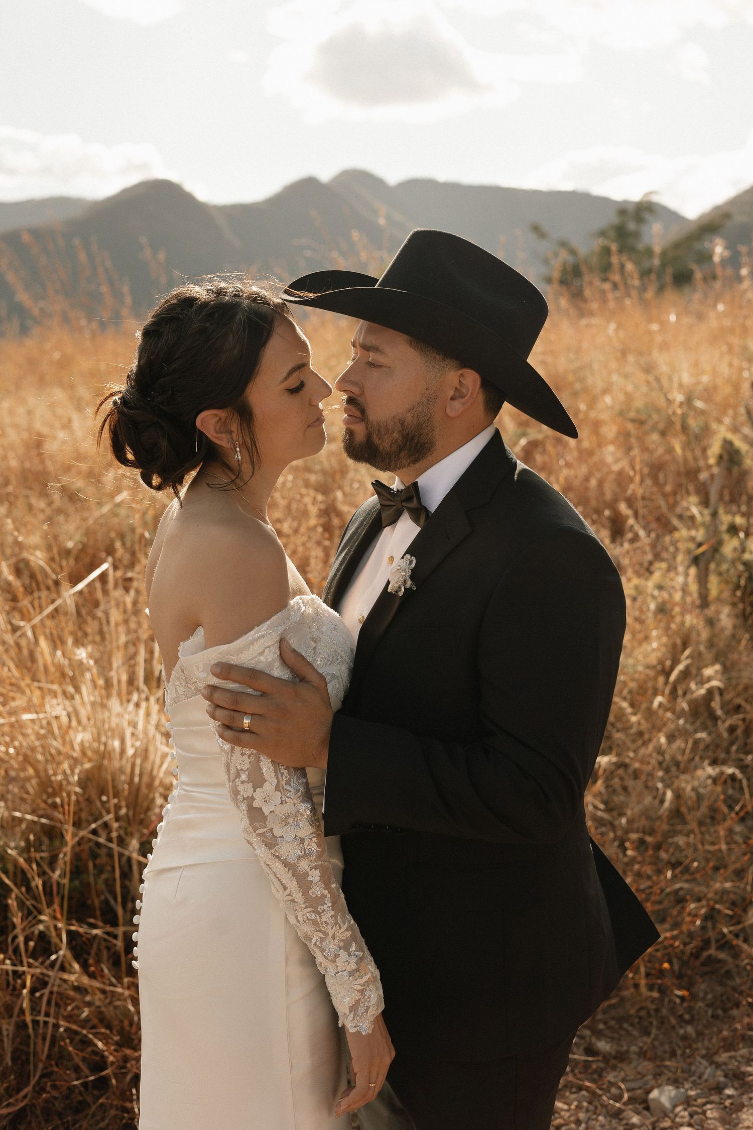 A couple embraces in a romantic golden hour photoshoot amidst tall grass and mountains in the background.