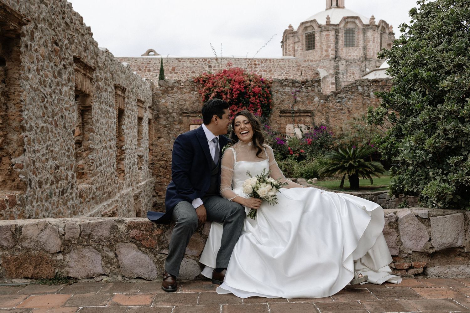 A couple in wedding attire sits together on ancient stone steps surrounded by historic stone walls and greenery.