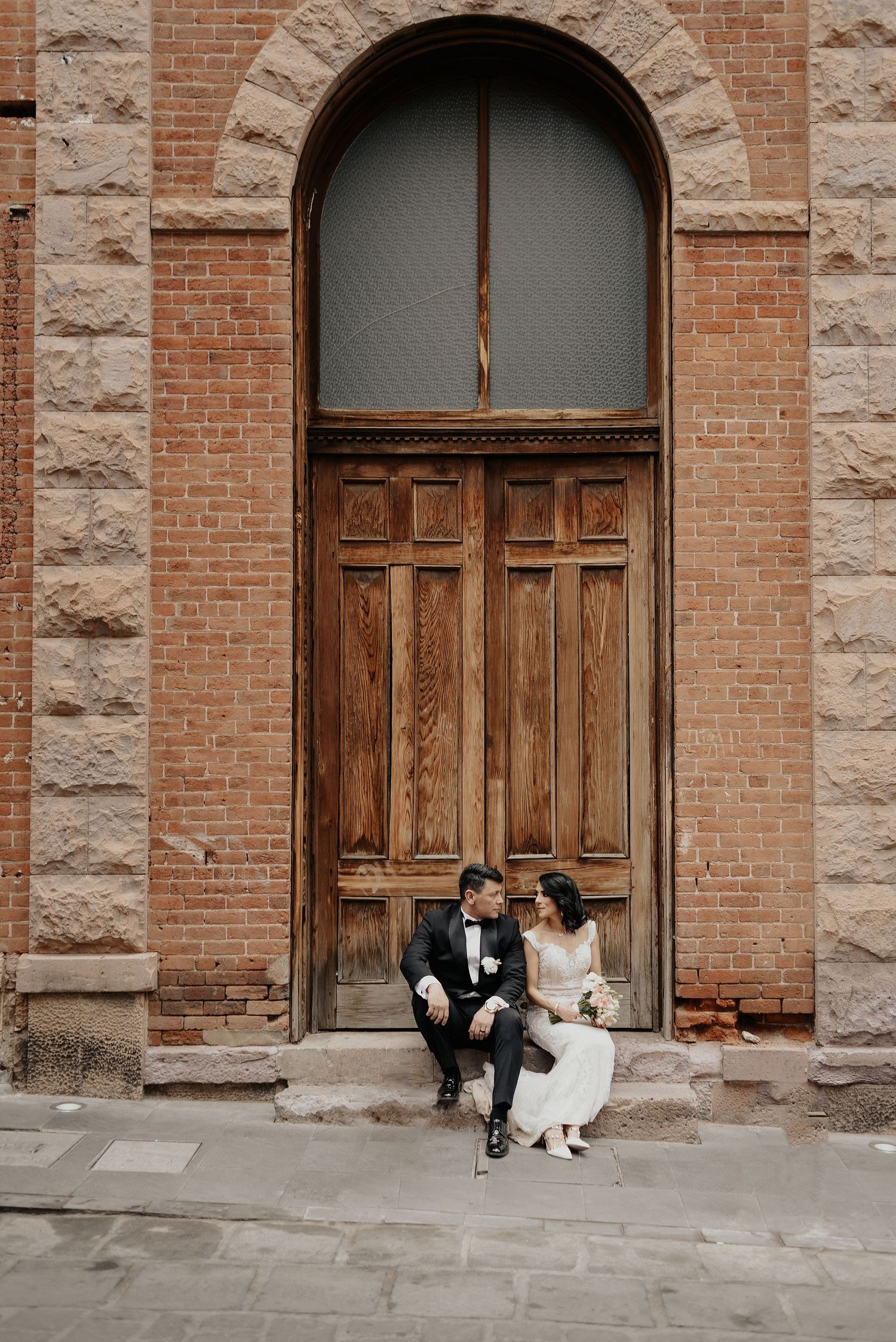 Wedding couple sitting beneath large arched wooden door in historic brick building.