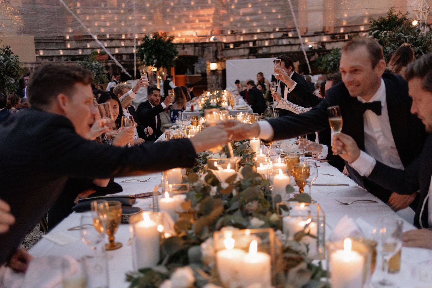 Wedding reception guests enjoying conversation at an elegantly decorated candlelit table with greenery centerpieces.