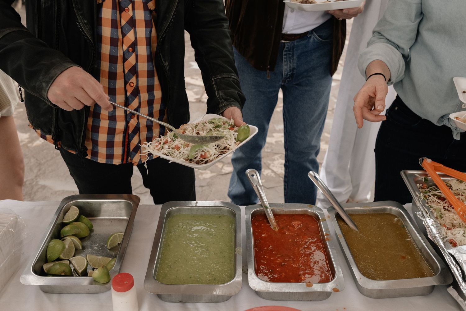 A buffet line shows various colorful dips and sauces in metal serving trays with people serving themselves.