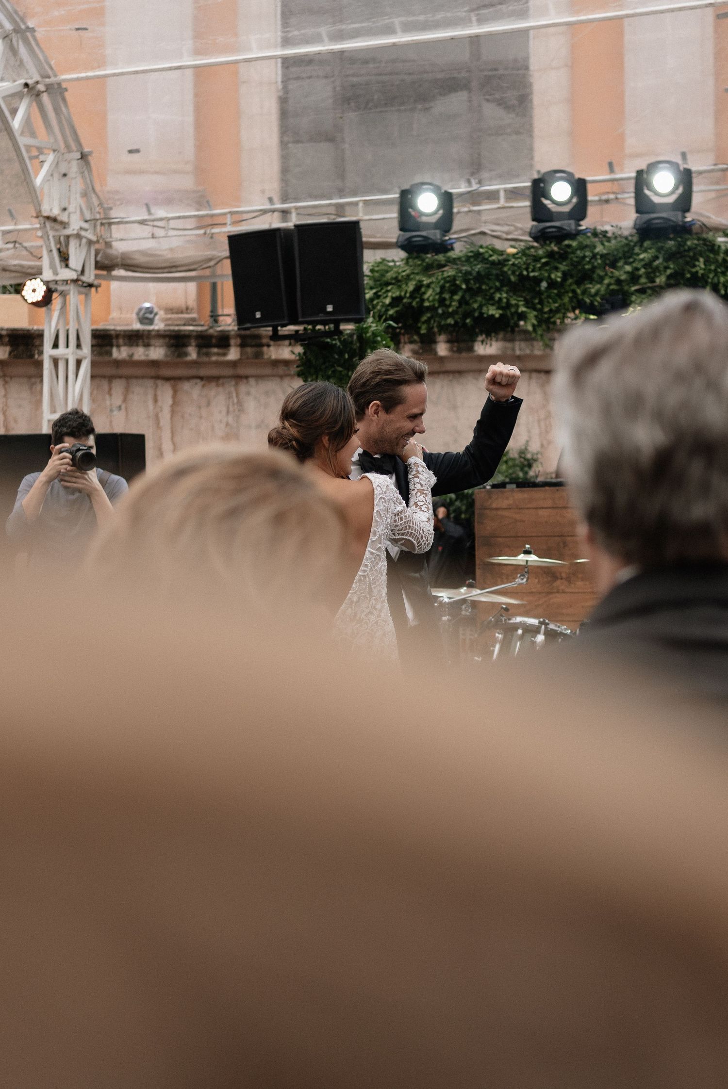 People gathered at an outdoor event with stage lighting and greenery visible against a peach-colored wall in the background.