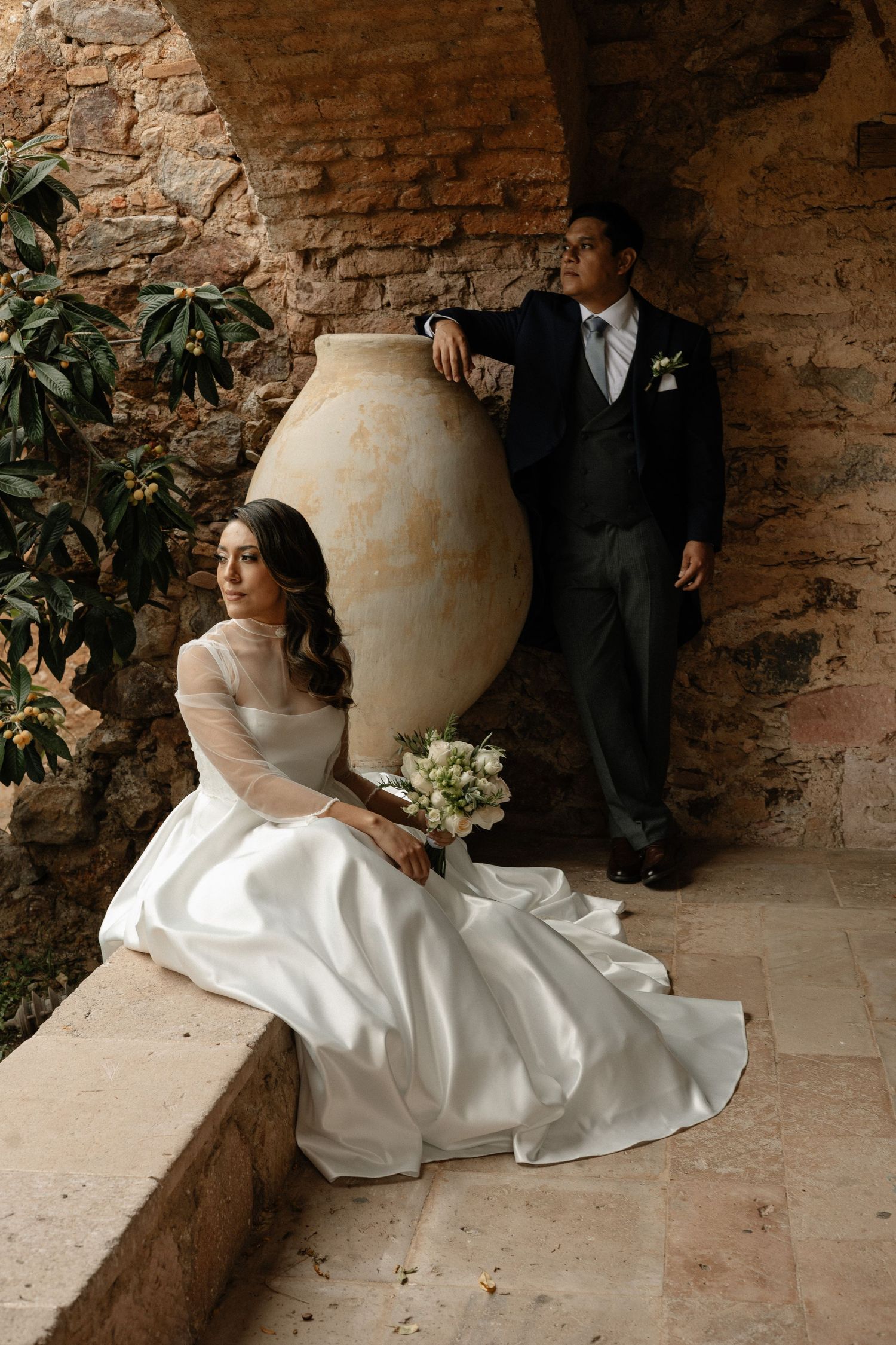 Newlyweds pose together against rustic brick wall with large decorative clay urn in romantic wedding portraits.