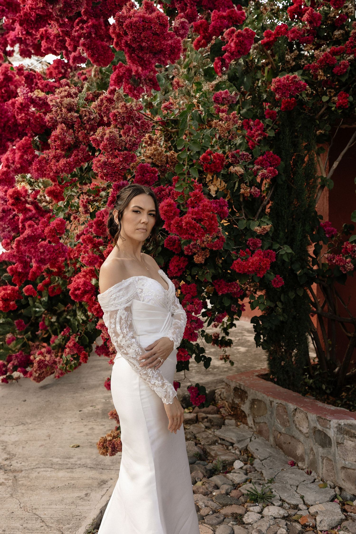 A white lace wedding dress is showcased against a vibrant backdrop of red bougainvillea flowers and stone wall.