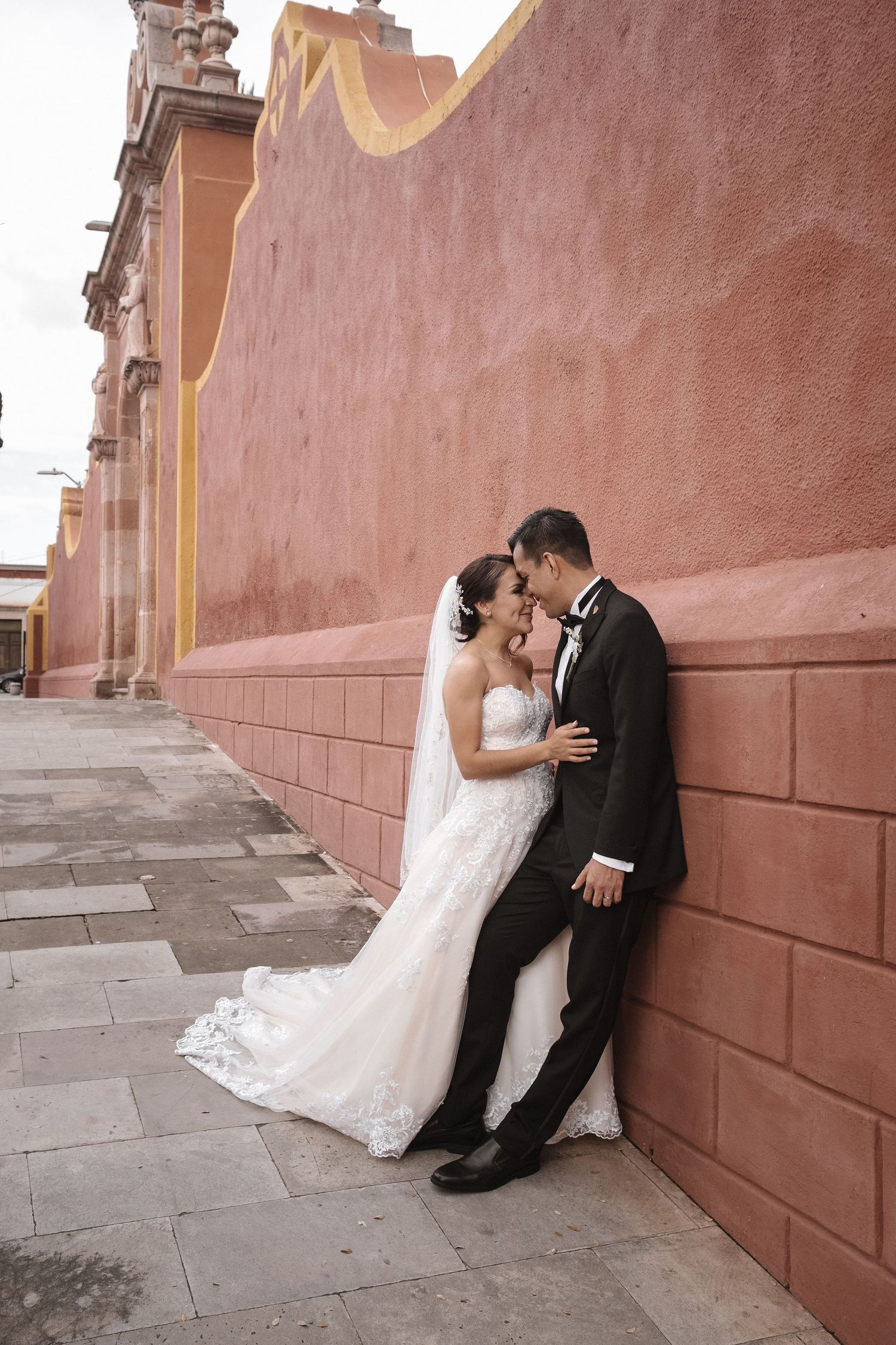 A romantic wedding photo against a terracotta wall with a flowing white dress and classic black tuxedo.