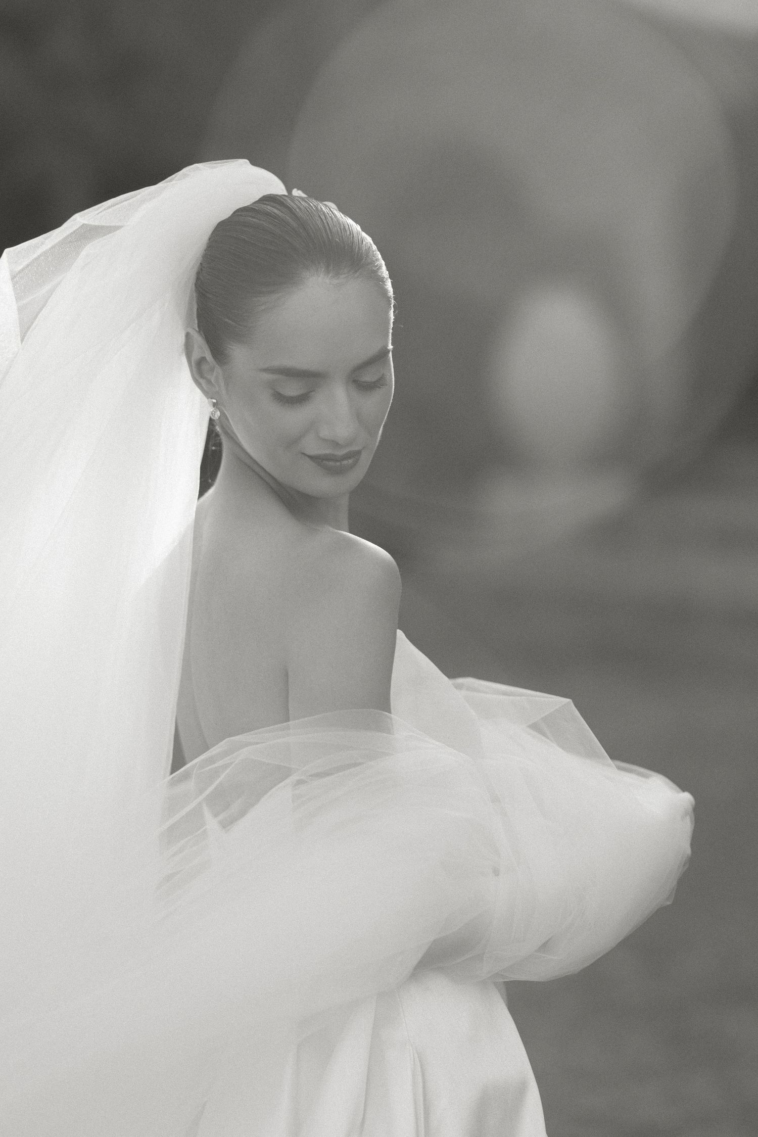 Black and white photo of a bride in a flowing white gown and veil, looking down gracefully outdoors.