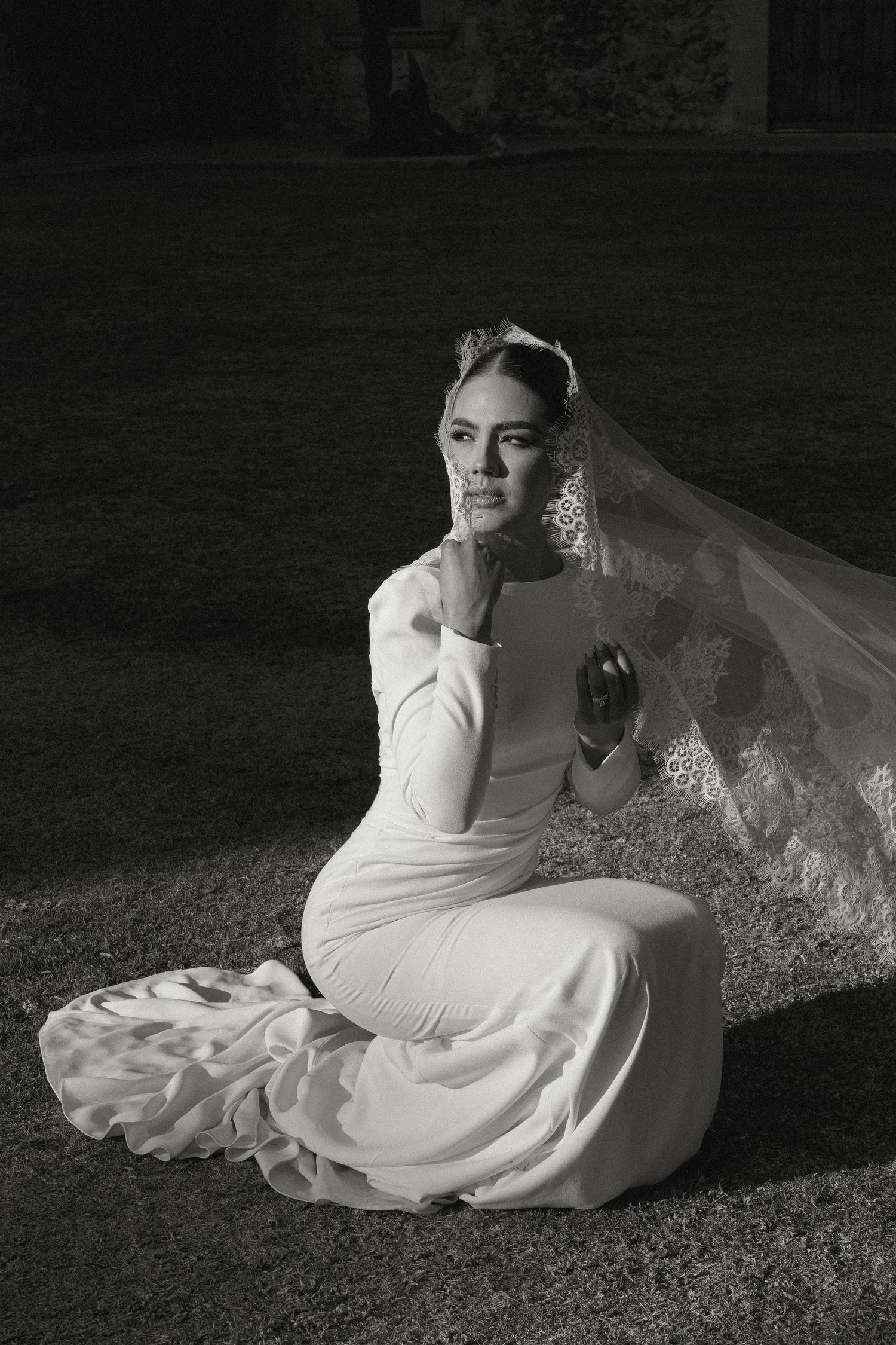 Black and white photo of a bride in a white gown and lace veil sitting gracefully on grass outdoors.
