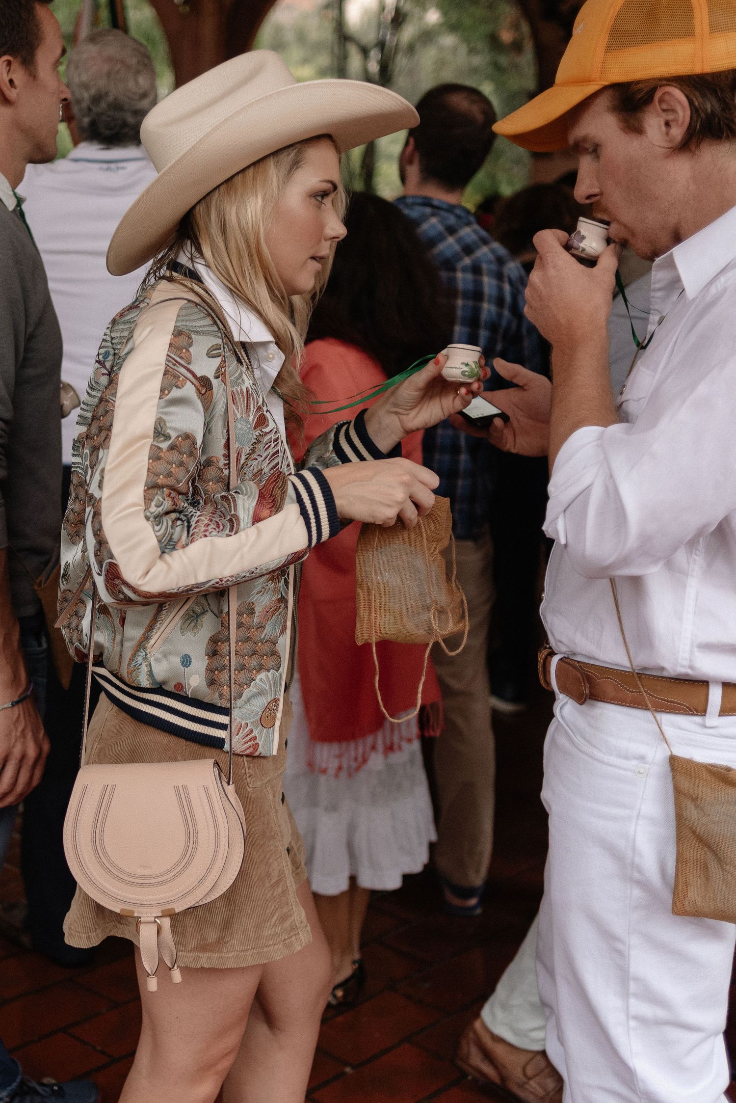 Two people in western attire with cowboy hats converse at a social gathering, one wearing a patterned jacket and tan skirt.