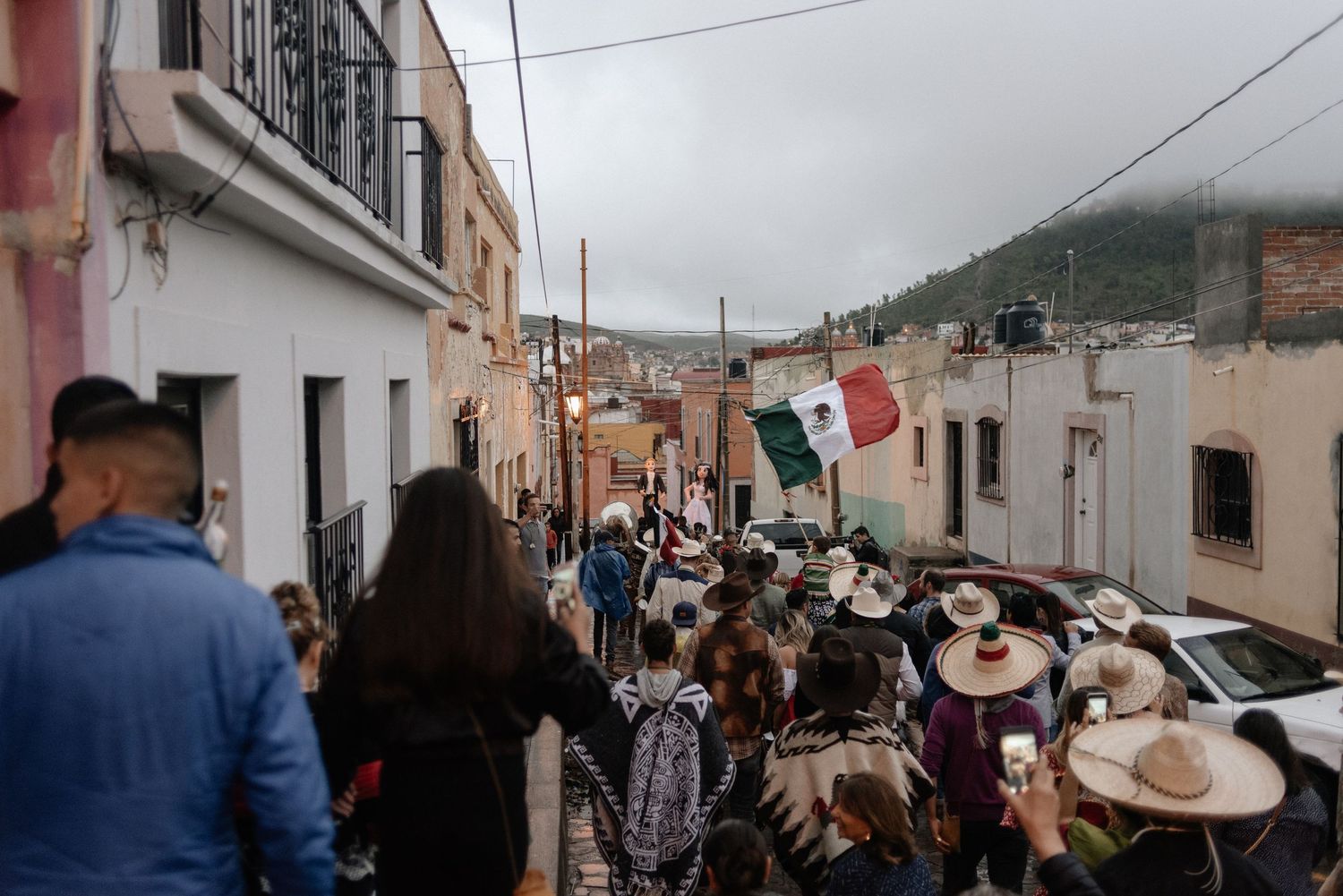 A crowded street scene in Mexico with traditional sombreros and flags visible between colonial-style buildings.