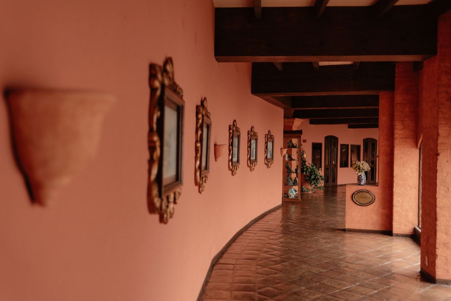 Curved pink hallway with wooden beams overhead and framed artwork lining the adobe-style walls.