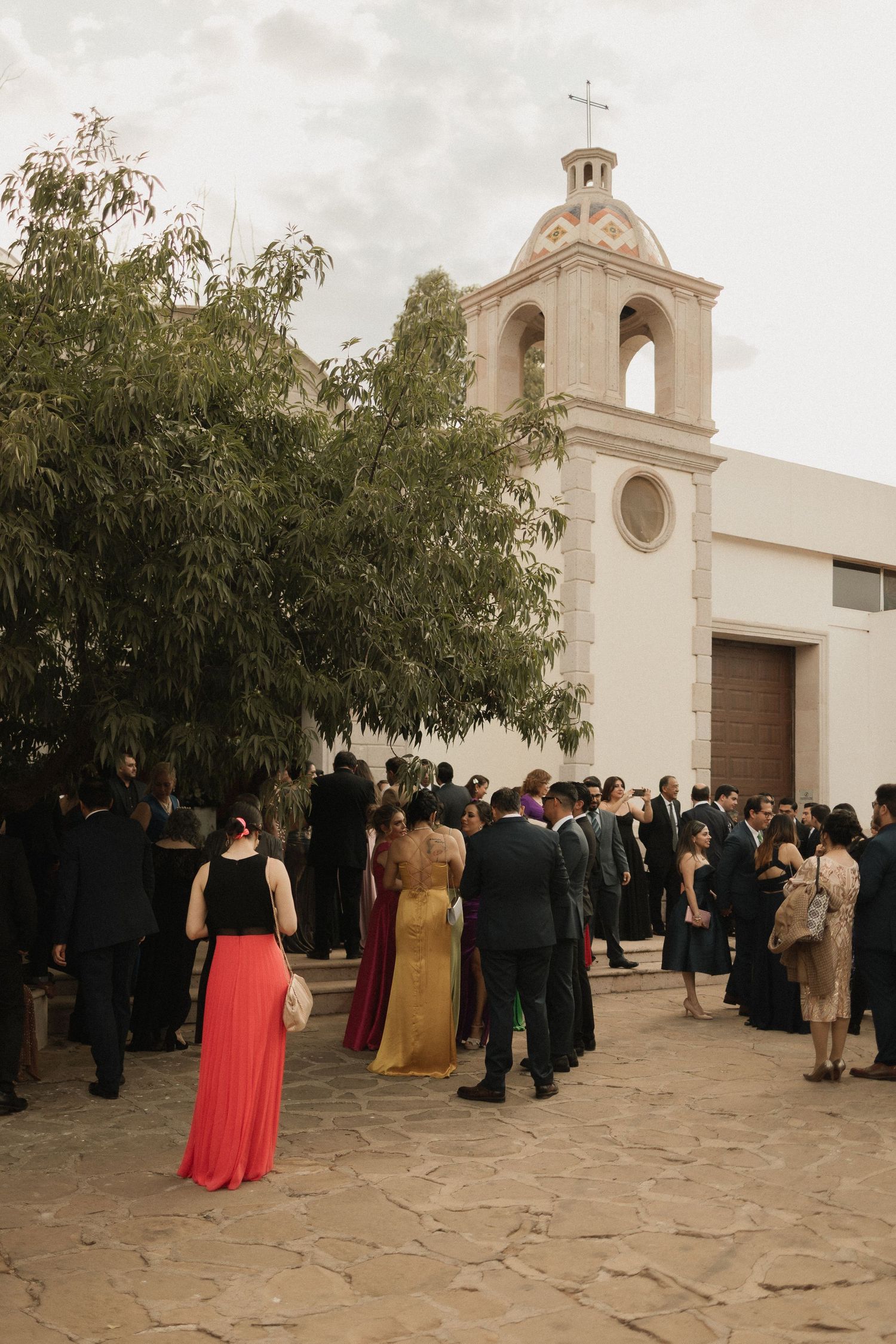 Wedding guests gather outside a white Spanish-style church with a bell tower during a celebration ceremony.