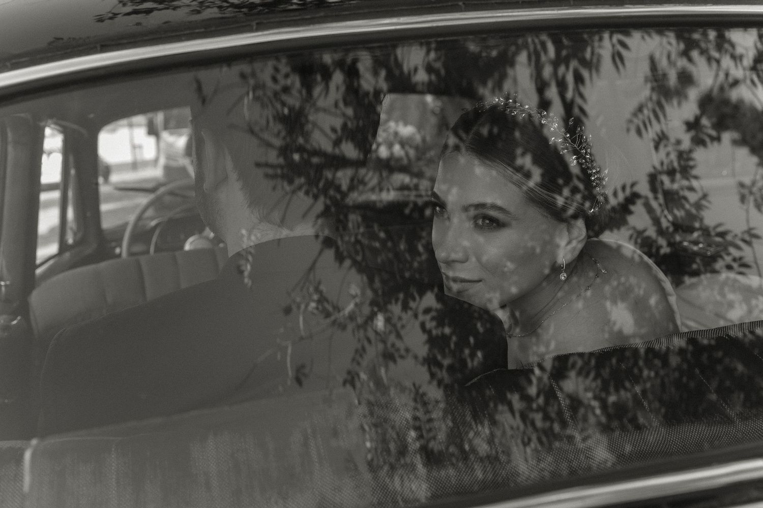 Black and white photo of trees reflected in a car window creating a moody silhouette effect.