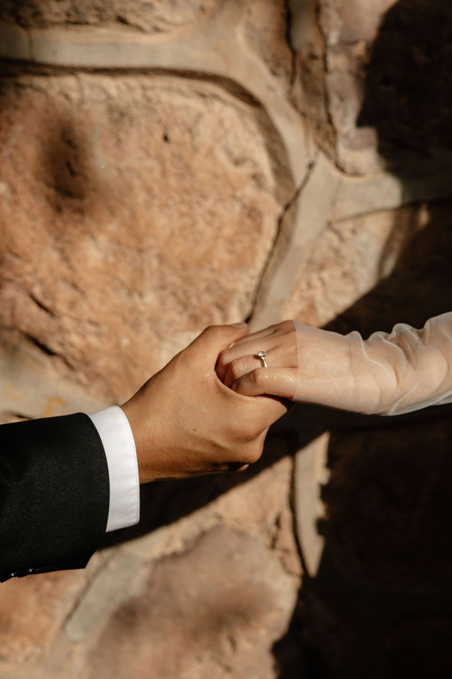 A close-up view of hands placing a wedding ring on a finger against a textured stone wall background.