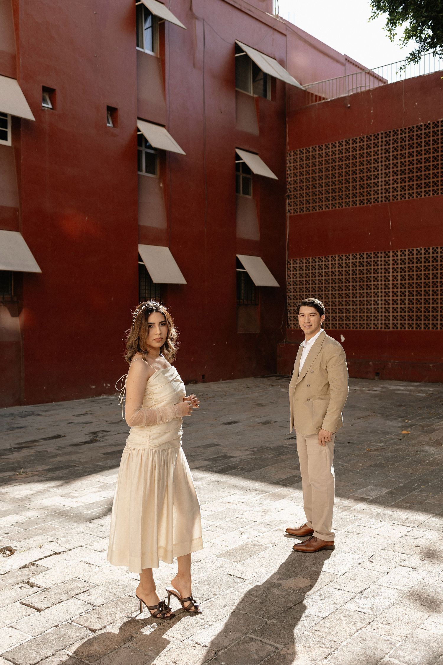 Two people in beige outfits pose against a red brick wall in an outdoor courtyard setting.