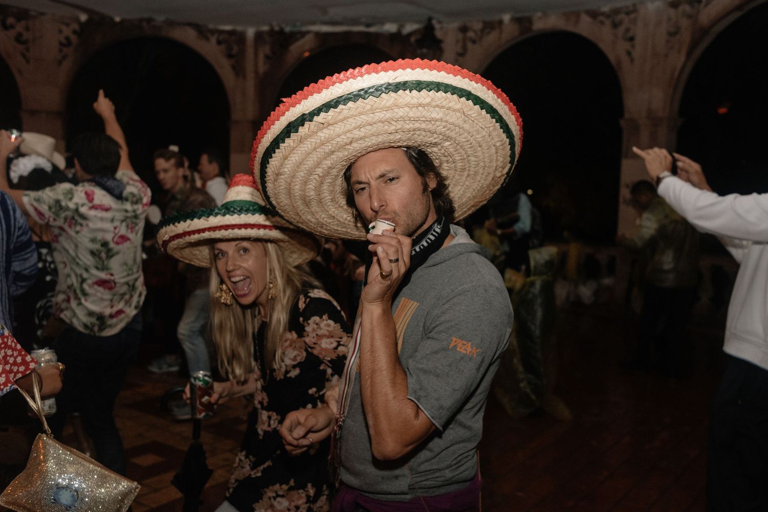 People enjoying a party wearing large Mexican sombreros in a dimly lit venue with arched architecture in the background.