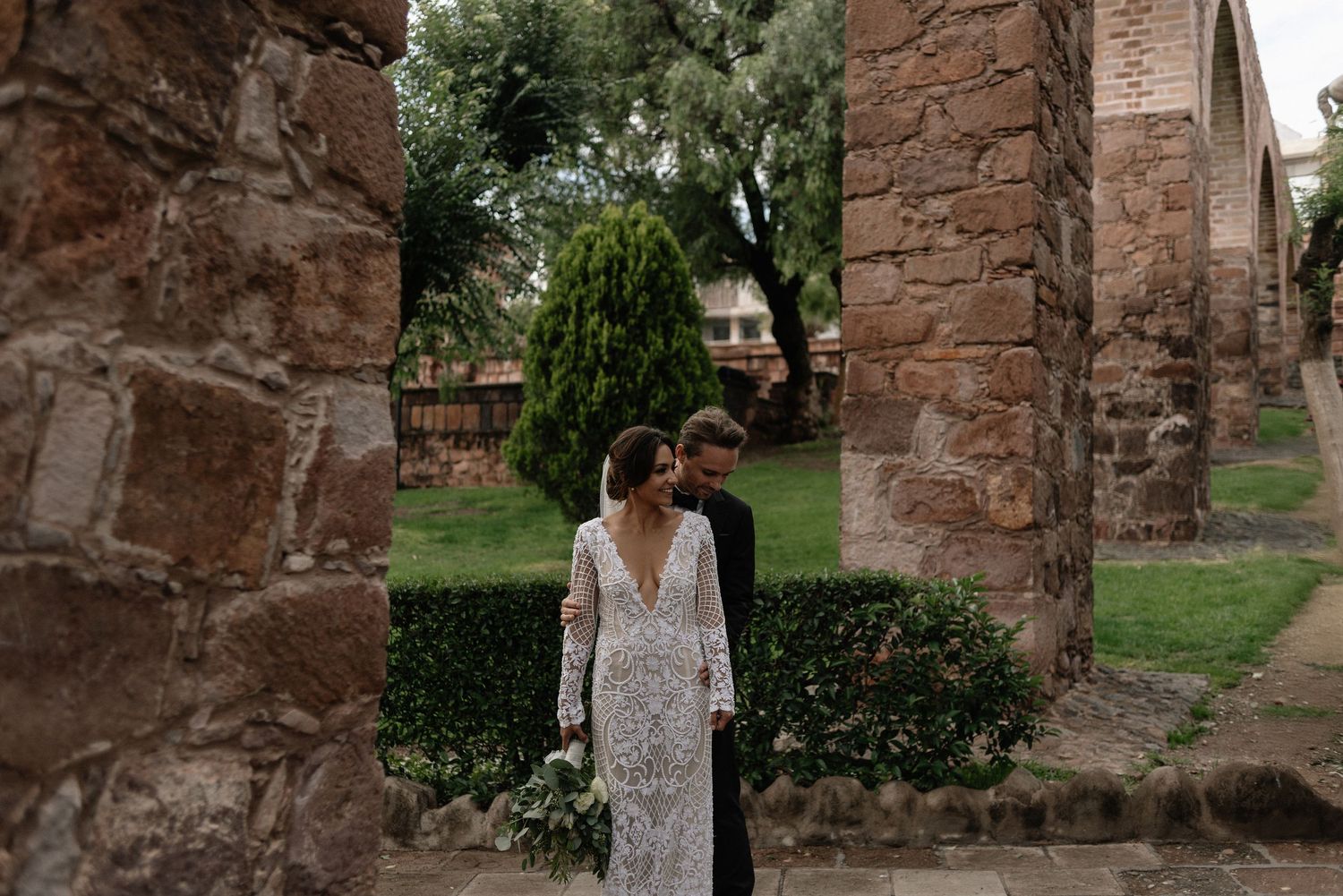 A lace wedding dress is showcased among stone archways and lush garden greenery at a historic venue.