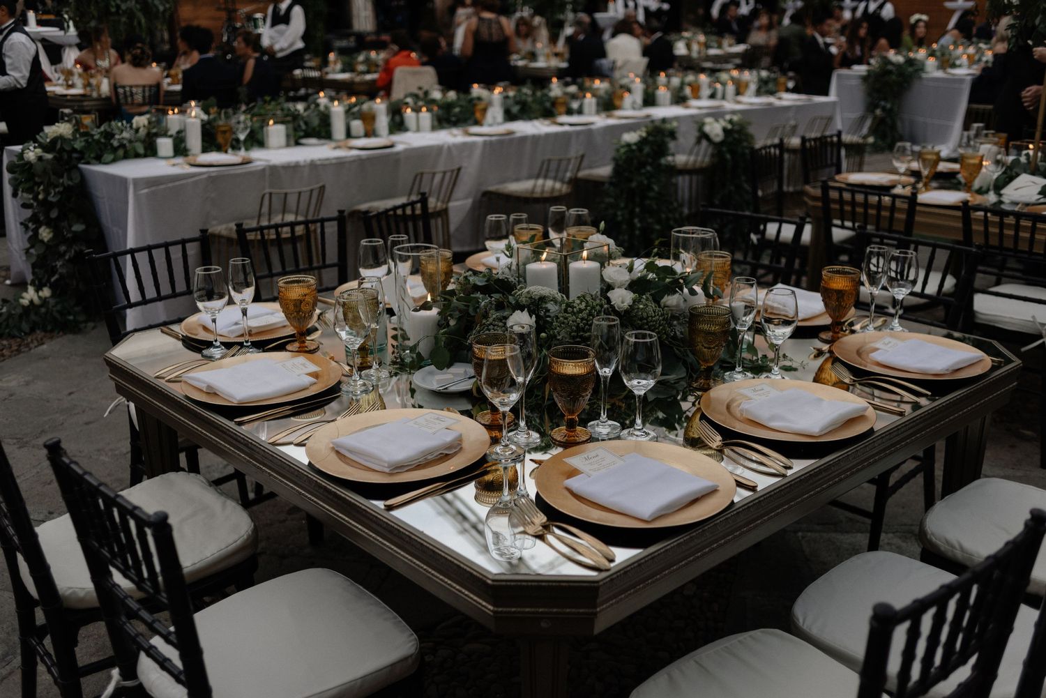 Elegantly set wedding reception tables with gold chargers, white plates and dark chairs in an indoor venue setting.