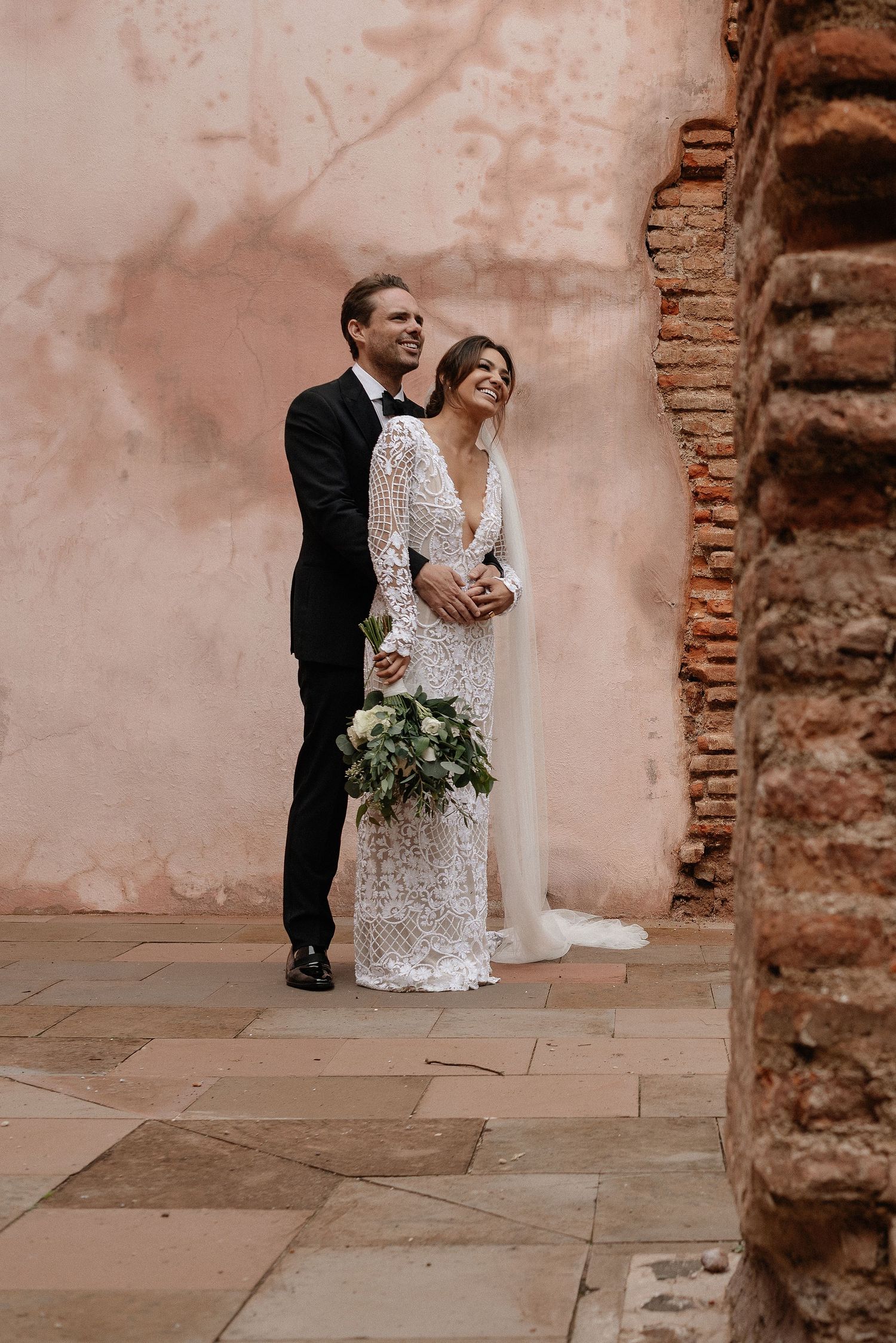 A couple in formal wedding attire embrace against an aged terracotta wall with exposed brick detail.