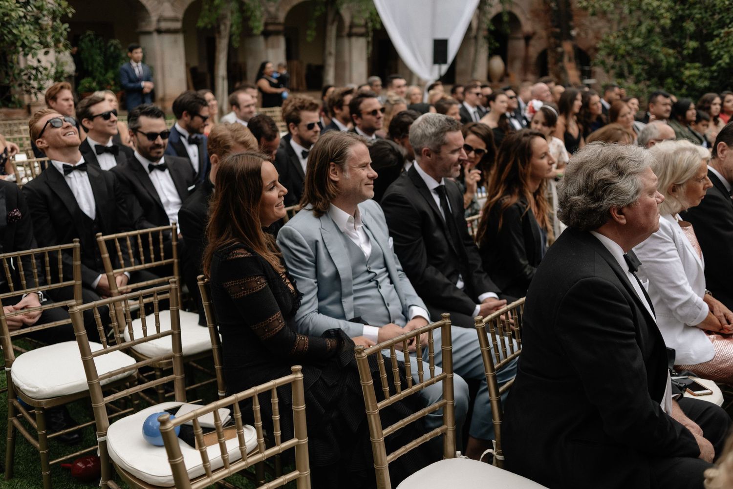 Outdoor wedding ceremony guests seated in gold chiavari chairs in a courtyard setting.