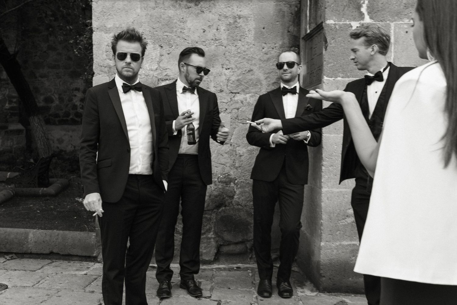 Four men in formal black tuxedos and sunglasses stand together holding drinks in vintage black and white photograph.