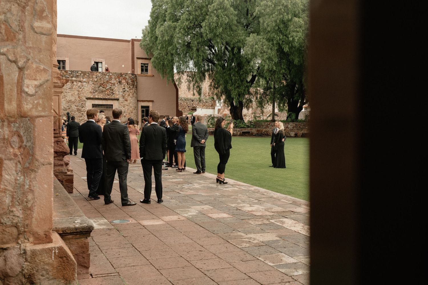 People walking along a stone path by a grassy courtyard next to historic brick buildings and trees.